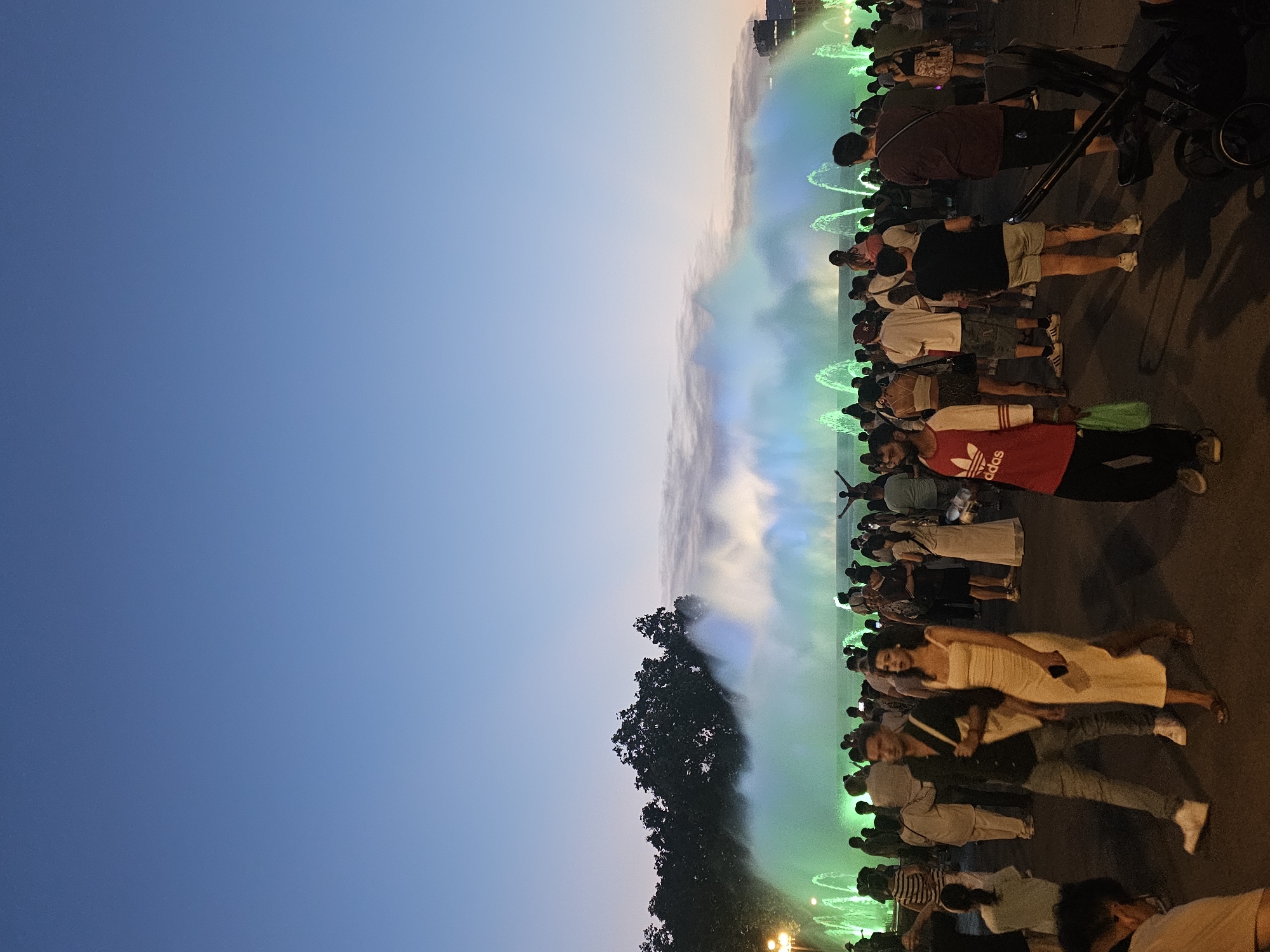 The image captures a group of people gathered in an outdoor setting, likely during an evening event. A water fountain display, illuminated with vibrant green and blue lights, dominates the right side of the frame, creating a misty, ethereal atmosphere. The sky transitions from a clear, deep blue at the top left to a lighter, warmer hue near the horizon on the right, suggesting either sunrise or sunset.
People are seen standing and watching the fountain show, some are dressed casually in t-shirts and shorts, while others are in dresses or more formal attire. A stroller is visible in the foreground, indicating families might be present. The ground appears to be asphalt, suggesting a public plaza or promenade. The overall mood seems to be one of shared enjoyment and wonder as the crowd observes the spectacle.
The lighting and the presence of the fountain suggest a public performance or attraction, possibly a tourist spot in Barcelona, Spain. The illumination from the fountain and some faint lights in the distance hint at it being dusk or early evening. There is no visible text in the image.