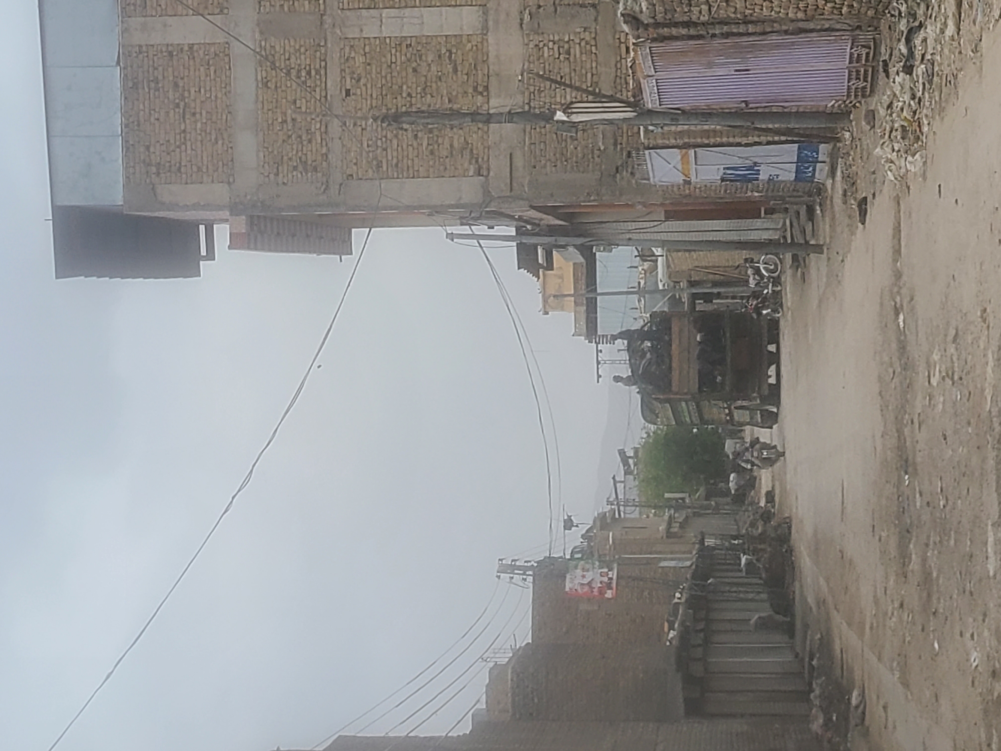 A street scene in Quetta City Tehsil, Pakistan is depicted. The street is unpaved with buildings on either side. The buildings are primarily constructed of brick and concrete, with some featuring corrugated metal roofing. Power lines stretch across the street, connecting to utility poles. A truck and motorcycles are visible along the street. The sky is overcast, creating a grey ambiance. The environment appears urban with visible signs of decay and litter.