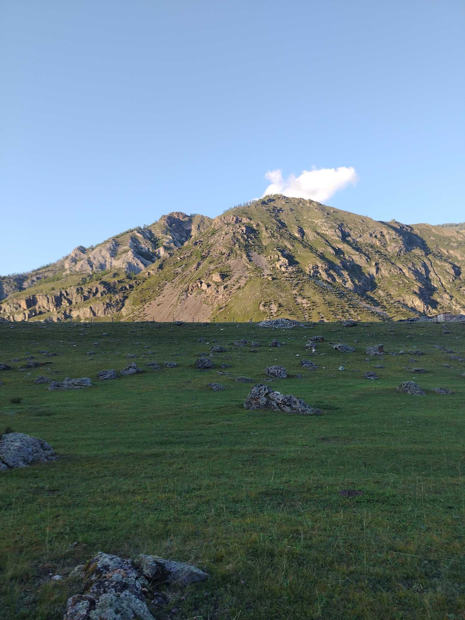 The image captures a scenic mountain landscape under a clear blue sky with a single white cloud. In the foreground, a vast green meadow is dotted with numerous grey rocks of various sizes. The middle ground reveals the lower slopes of a rugged mountain range, characterized by steep, rocky formations and patches of green vegetation, with some sparse trees visible higher up. In the far distance, a few buildings can be seen nestled at the base of the mountains. The lighting suggests it is either early morning or late afternoon, casting long shadows across the mountain slopes. The location is identified as Iodro, Russia.