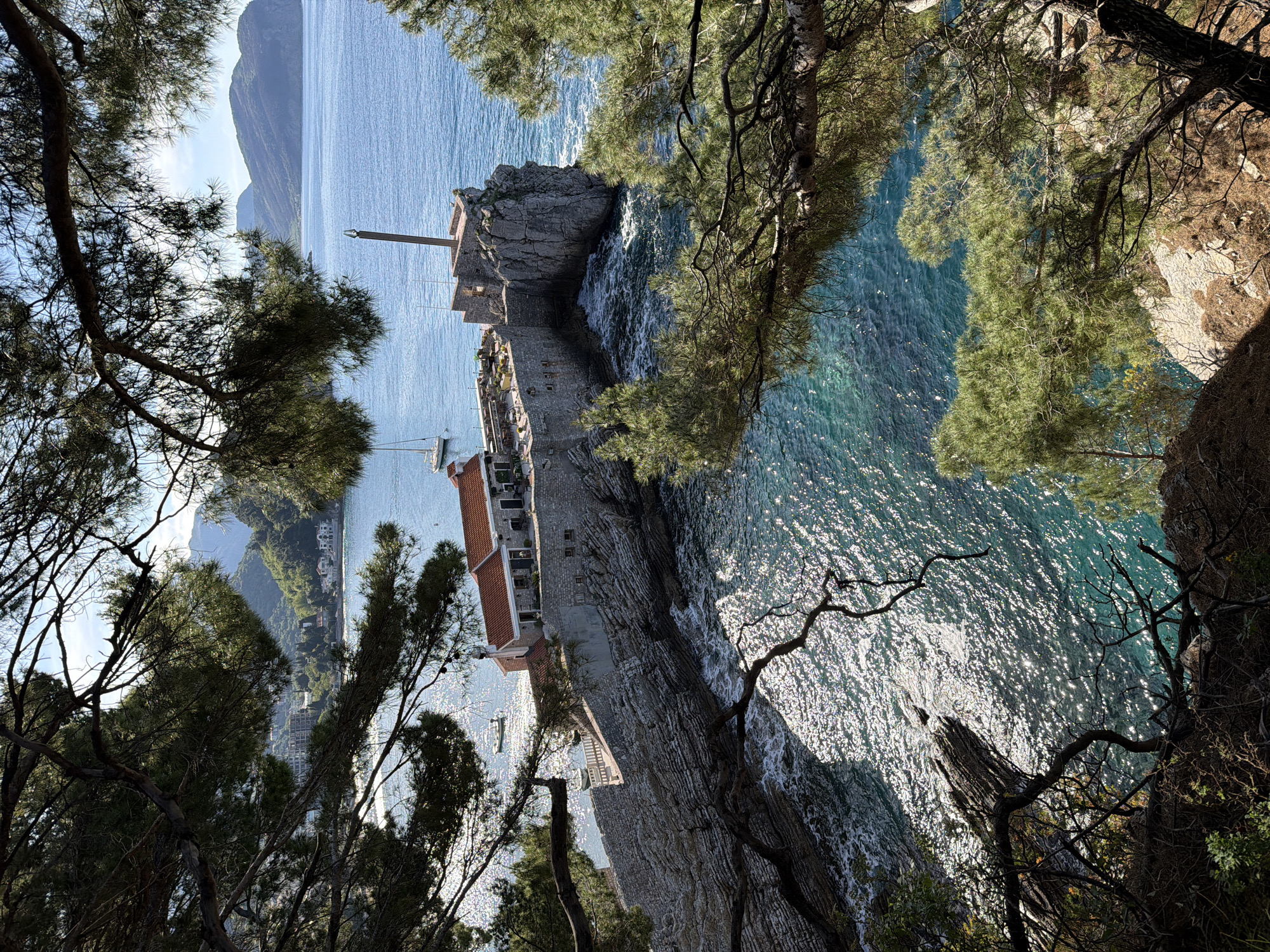 An elevated, framed view showcases a picturesque coastal scene, likely Petrovac, Montenegro. In the foreground, dark silhouettes of tree branches and foliage partially obscure the top, bottom, and left of the frame, suggesting the vantage point is from a wooded area.
The midground features an old stone fortress or coastal town built along a rocky cliff edge, extending into the clear, sparkling turquoise-blue sea. Buildings with distinctive red-tiled roofs are visible within and behind the fortifications. A tall, slender mast-like structure, possibly a flagpole or monument, stands prominently on a fortified outcrop. Several small boats, including at least two sailboats, are anchored or navigating the calm waters near the complex. White foam indicates where the sea meets the rocky shore.
The bright daylight causes intense glittering reflections on the water's surface, suggesting a sunny day, likely mid-morning or afternoon. In the background, the vast expanse of the sea stretches towards distant, hazy mountains under a clear sky. Some additional coastal development can be discerned at the base of these mountains. No people are visible, and there is no discernible text within the image.