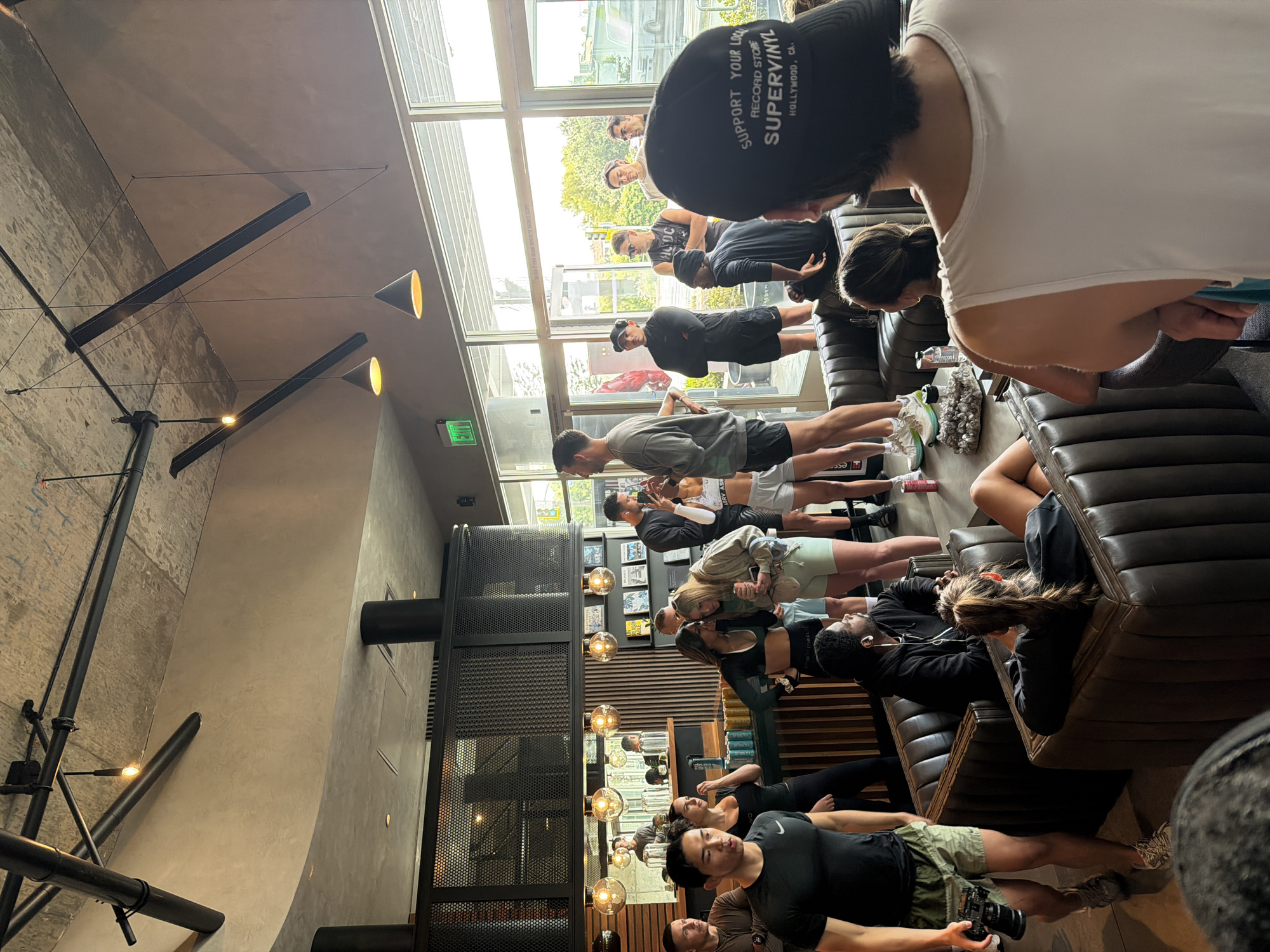 A medium shot captures a diverse group of people engaging in what seems to be a yoga or stretching session indoors, possibly in a studio or a large room with a cafe-like ambiance. The space is well-lit by both natural light streaming in from a large window and artificial lights hanging from the ceiling. The decor includes industrial-style black metal fixtures and beams, contrasting with the polished concrete walls.
The people in the group are diverse in age and ethnicity, dressed in athletic wear. They are arranged in a line, indicating a class or guided session. Some are focused on their exercises, while others appear to be interacting with each other.
The outdoor view visible through the window suggests a cityscape, hinting at a location within an urban setting. The presence of a "SUPPORT YOUR LOCAL RECORD STORE SUPERVINYL HOLLYWOOD, CA" cap indicates the location is likely West Hollywood, United States of America.