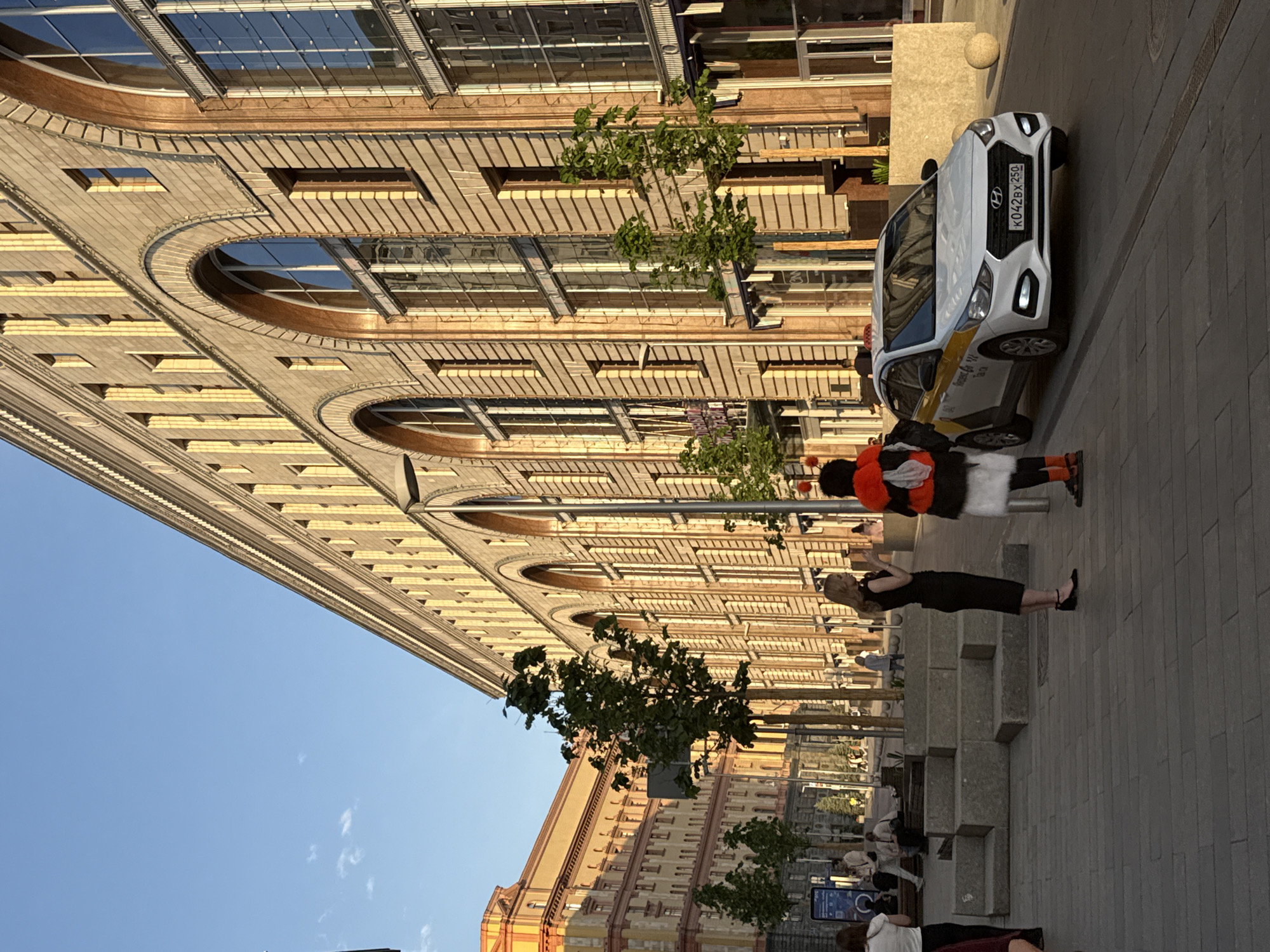 The image depicts a Moscow street scene, Russia, at ground level, rotated sideways. A white Hyundai Solaris taxi with license plate K042BX250 is parked on the right side of the image. A woman in a black dress and heels is standing on the paved sidewalk, seemingly in conversation with an individual dressed in a clown-like costume with a red and orange wig. The sidewalk is equipped with modern concrete seating and young trees. In the background, numerous people are seated on the ground. In the background are two large multi-story buildings, designed with repeating arched windows. The sky is clear and blue.