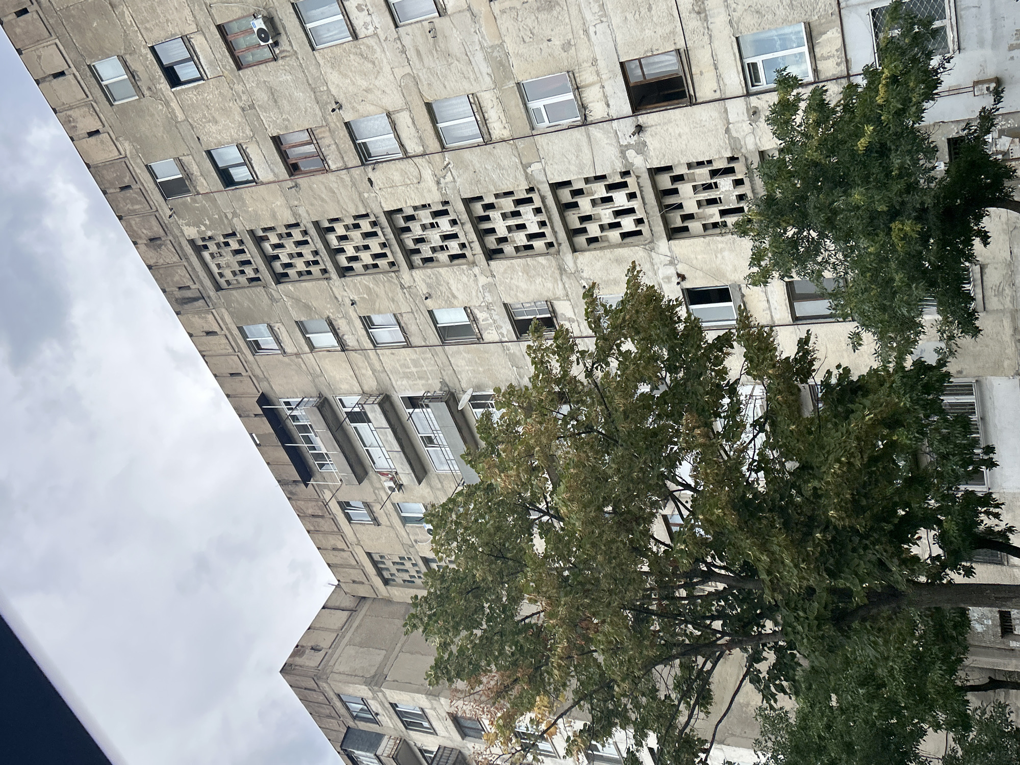 The image shows a Brutalist-style apartment building in Tbilisi, Georgia. The building is made of concrete and has a weathered facade. There are many windows and balconies, some of which are enclosed. The building has a unique architectural feature of vertical panels with patterned openings. In the foreground, there are trees with green and yellow leaves, indicating it is autumn. The sky is overcast. The image does not show any people or indicate any actions or interactions.
