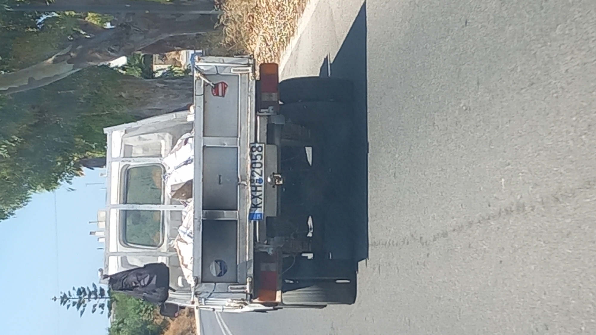 A rear view of a white truck with a metal flatbed is captured on a paved road in the city of Kos, Greece. The truck is loaded with several large, white sacks. A black backpack is hanging from the top of the truck's cab on the driver's side. The truck's license plate, visible on the rear, reads "K X H 2058". The background consists of dry vegetation, trees, and a clear blue sky, indicating a warm, sunny day. The truck is positioned in the right lane of the road, which appears to be a two-lane road. The scene suggests the truck is in motion, driving on a road on the outskirts of the city.
