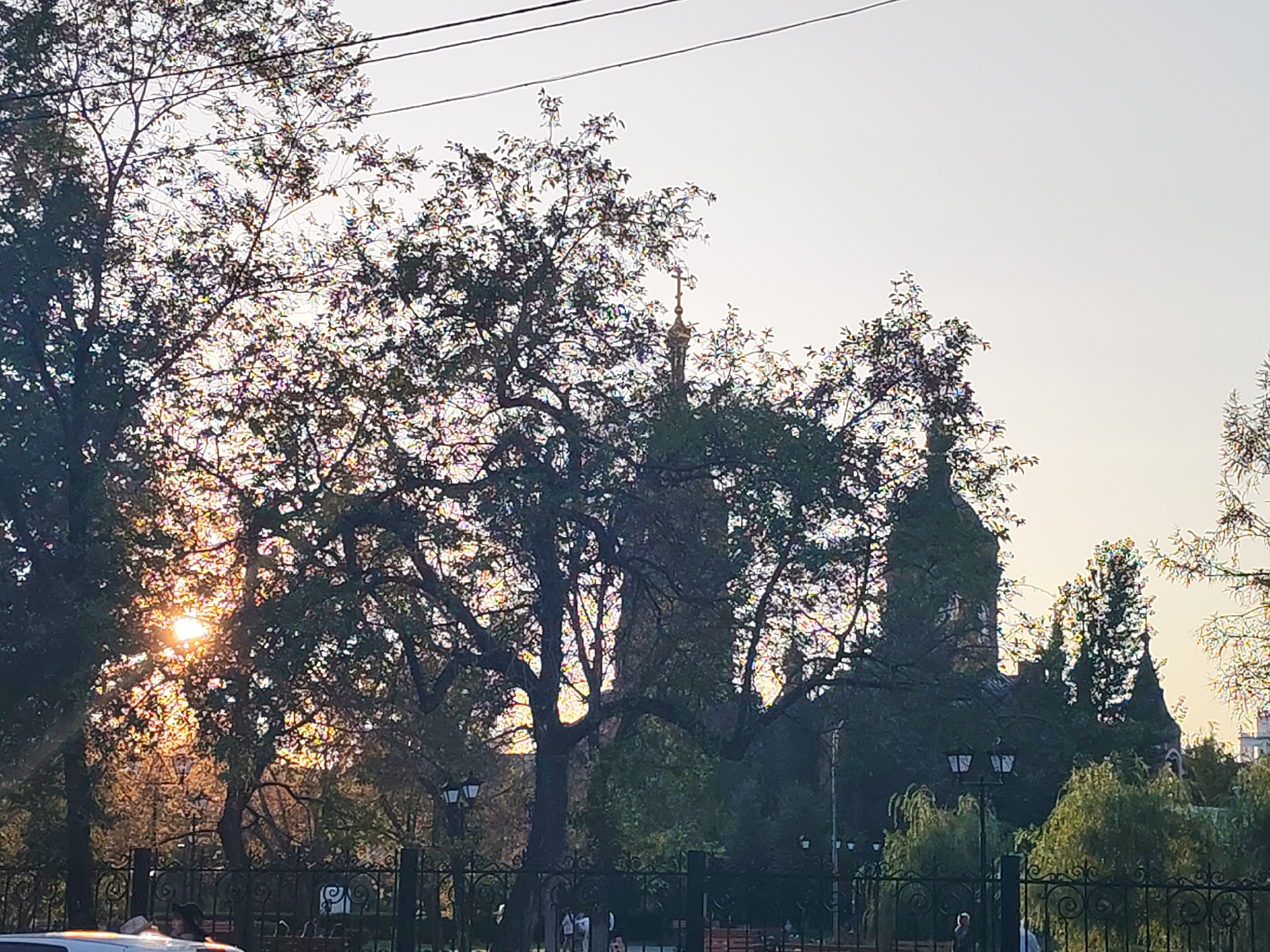 The image shows a park scene with trees and architecture in the background. Sunlight filters through the leaves of several trees in the foreground, creating a dappled light effect and a rainbow shimmer on the foliage. In the mid-ground, behind the trees and a black wrought-iron fence, are buildings. One building appears to be a church or cathedral with a tall steeple topped with a cross. Another structure, to the right, has several pointed turrets, partially obscured by trees. There are lampposts visible in the park. In the far background, the sky is a pale yellow-orange, indicating either sunrise or sunset. A few people are faintly visible in the background, some standing near the fence and others walking within the park. Part of a white car is visible in the lower left corner of the image. The overall impression is that of an urban park setting, possibly with historical buildings.