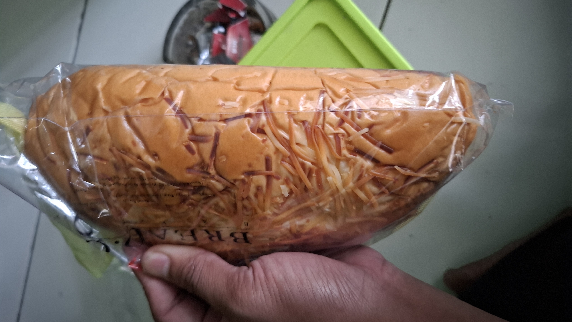 A person's hand, with skin tone appearing medium-dark, holds a loaf of bread that is packaged in clear plastic. The bread is golden brown and appears to be topped with shredded cheese, some of which is visible on the surface. The bread itself has a soft, yielding texture, evident from the indentations on its surface. The packaging is sealed with a yellow and green label.
The background consists of a tiled floor, a bright green plastic container, and a blurry glimpse of a glass container with red items inside. This scene appears to be indoors, likely in a home or a small food establishment in Cilaku, Indonesia. The lighting is neutral, suggesting it is daytime. There are no other individuals clearly visible in the frame.