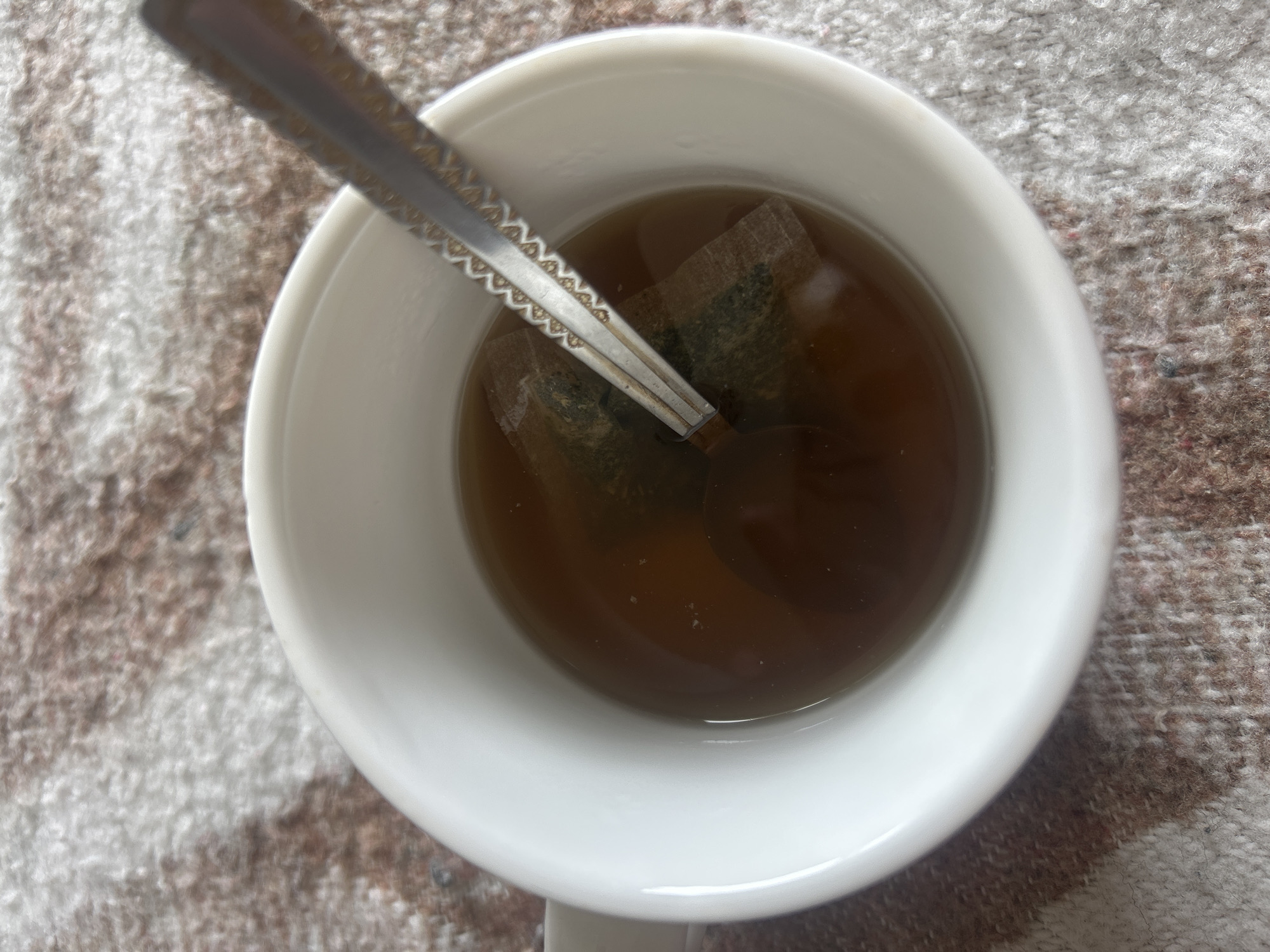 A top-down shot shows a white mug filled with dark brown liquid, presumably tea, with two tea bags steeping inside. A silver spoon rests in the mug. The mug is set on a textured, patterned surface that resembles a brown and white rug. The scene is indoors, with details limited to the mug, its contents, the spoon, and the immediately surrounding surface. There is no indication of location in Burgas, Bulgaria or any other city. No people or interactions are depicted.