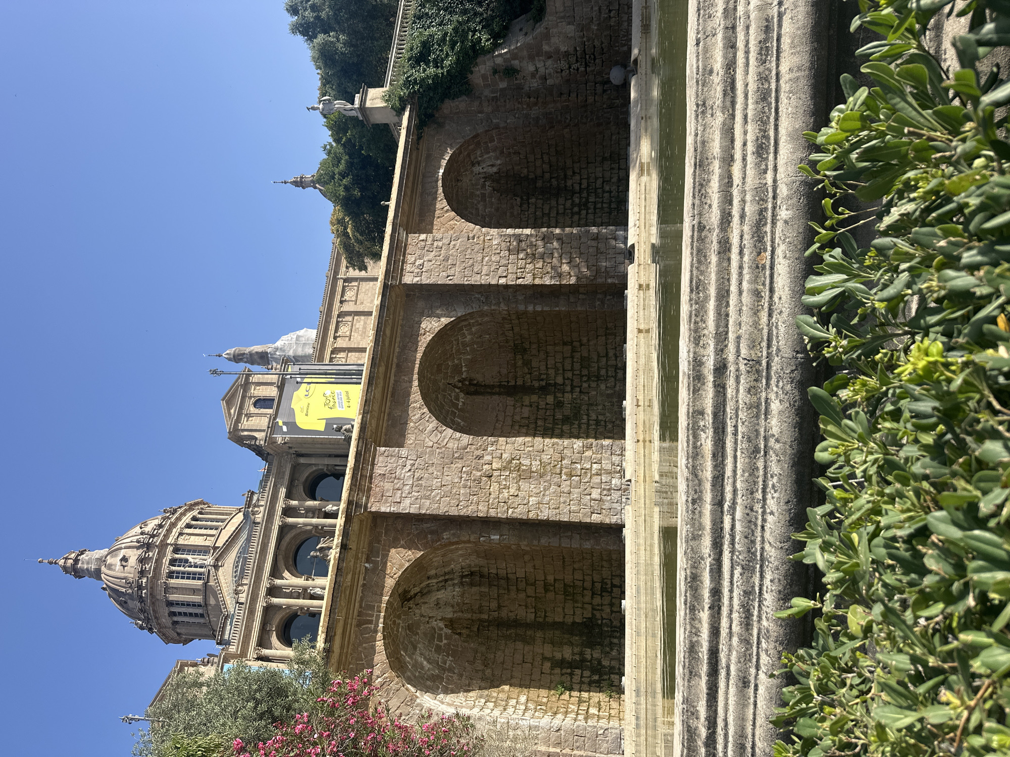 This is a photograph of the Palau Nacional in Barcelona, Spain, a grand building with a prominent dome and classical architecture. In the foreground, a tiered fountain with calm water is visible, surrounded by lush green foliage. Behind the fountain is a stone wall with arched alcoves, likely part of the building's elaborate facade. Above the wall, the main structure of the Palau Nacional rises, featuring columns, windows, and sculptures. A bright yellow banner with text is displayed on the building. The sky is clear blue, indicating a sunny day. There are no people visible in the frame. The scene conveys a sense of historical grandeur and public space.