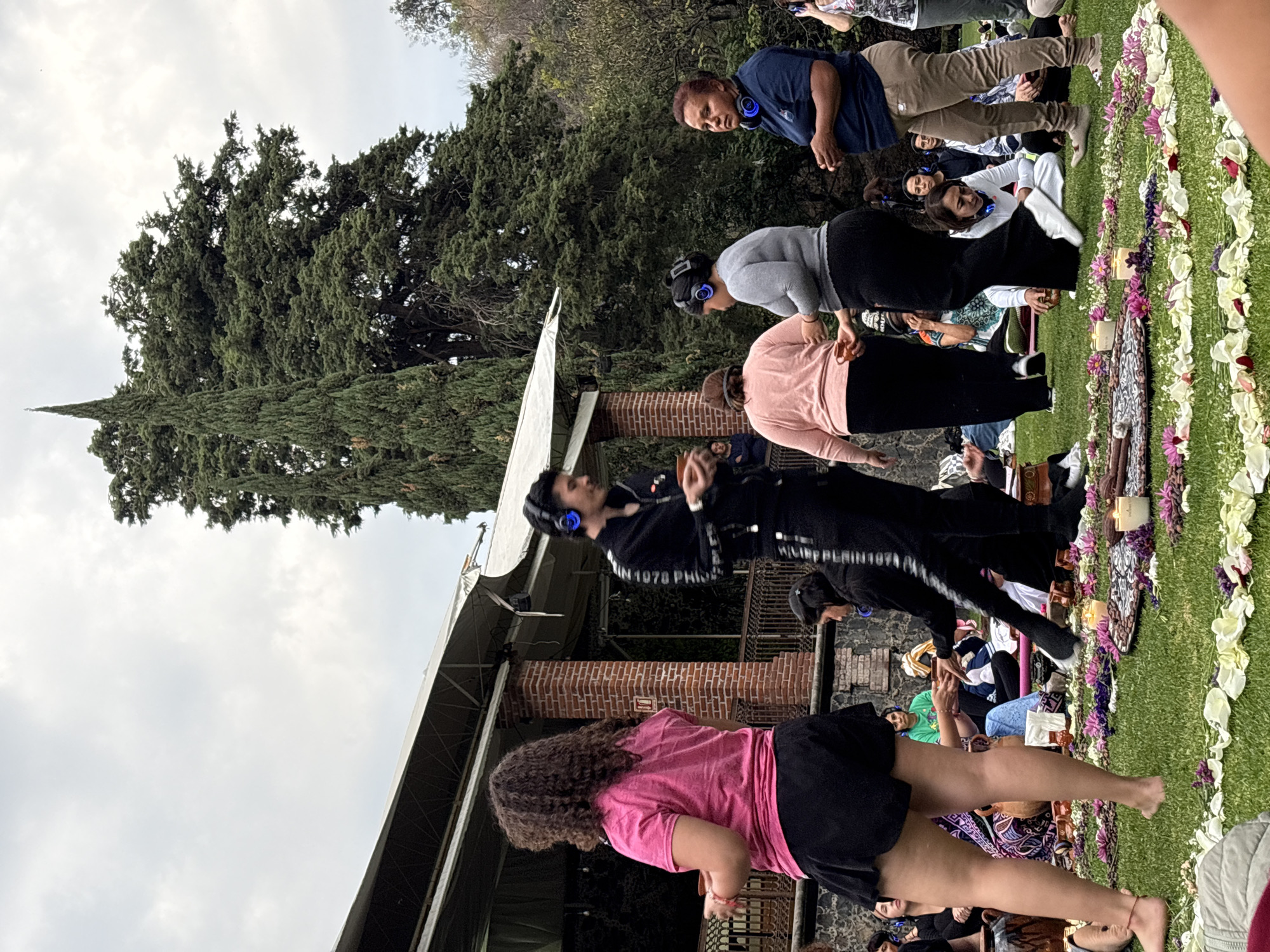 A scene in Santa Ana Acozautla, Mexico, shows a group of people gathered outdoors, potentially for a silent disco or similar event. They are in a grassy area next to a low building with brick pillars and a canopy. Tall trees are visible in the background. People are standing and moving, many wearing headphones. A decorative floral arrangement, possibly for an event, is on the grass. The sky is overcast.