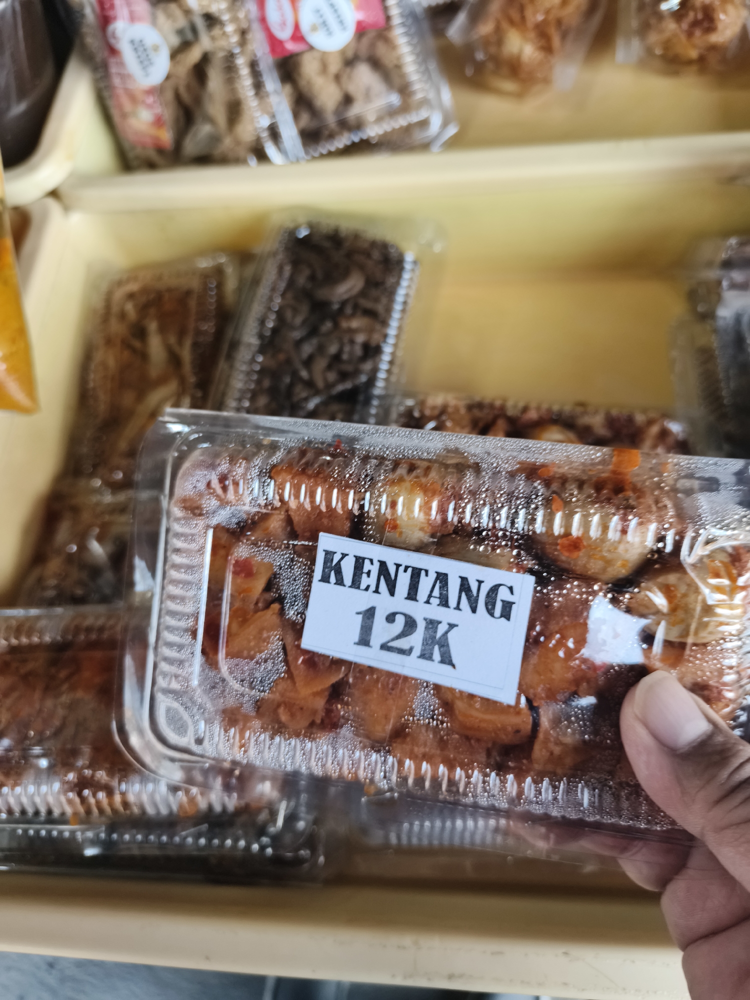 In a market stall in Petiken, Indonesia, an assortment of packaged snacks is displayed. In the foreground, a clear plastic container holds fried potato wedges, labeled with a white sticker that reads "KENTANG 12K". The container is condensation-covered, indicating it might have been refrigerated or is fresh. Behind it, other clear plastic containers are visible, some filled with dark, dried food items, possibly small fish or insects, and others with what appear to be larger, fried snacks. The snacks are arranged in a wooden or plastic tray. The background is softly blurred, showing more packaged goods. A hand is visible on the right side of the frame, holding the container of potato wedges.
