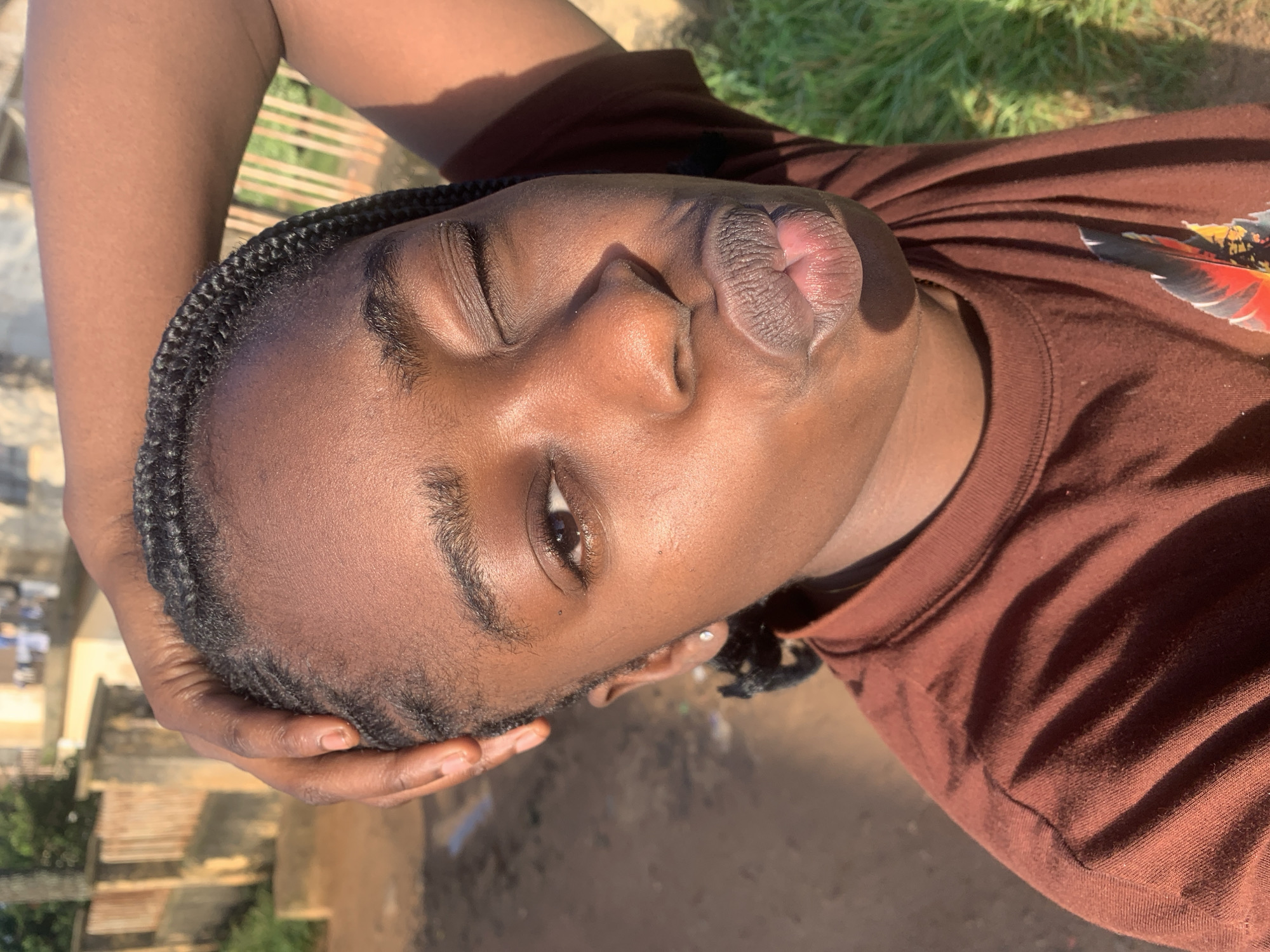 In Rafin Par, Nigeria, a close-up shot captures a young Black female from the shoulders up, outdoors during daytime. She is wearing a brown t-shirt and her left arm is raised, with her hand resting on her braided hair. Her head is tilted slightly back, and she is winking while puckering her lips. The background shows a dirt path and some vegetation, with a building and fence visible in the distance. The lighting suggests direct sunlight.