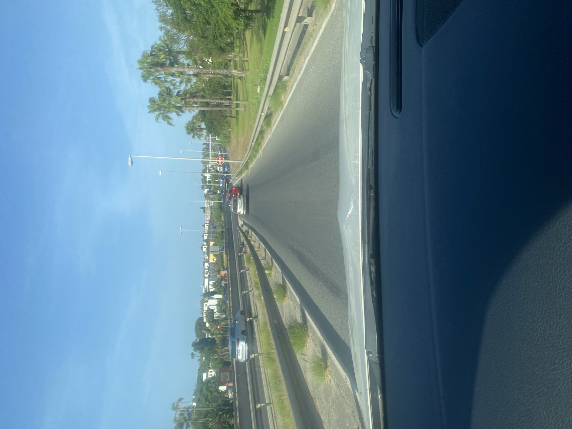 The image shows a view from inside a vehicle driving on a highway in Estepona, Spain. The highway has multiple lanes and is bordered by a metal guardrail and a grassy median. Palm trees and buildings are visible in the background, suggesting a Mediterranean climate. Several cars are driving on the highway. The sky is clear and blue. The dashboard of the vehicle is visible in the foreground.