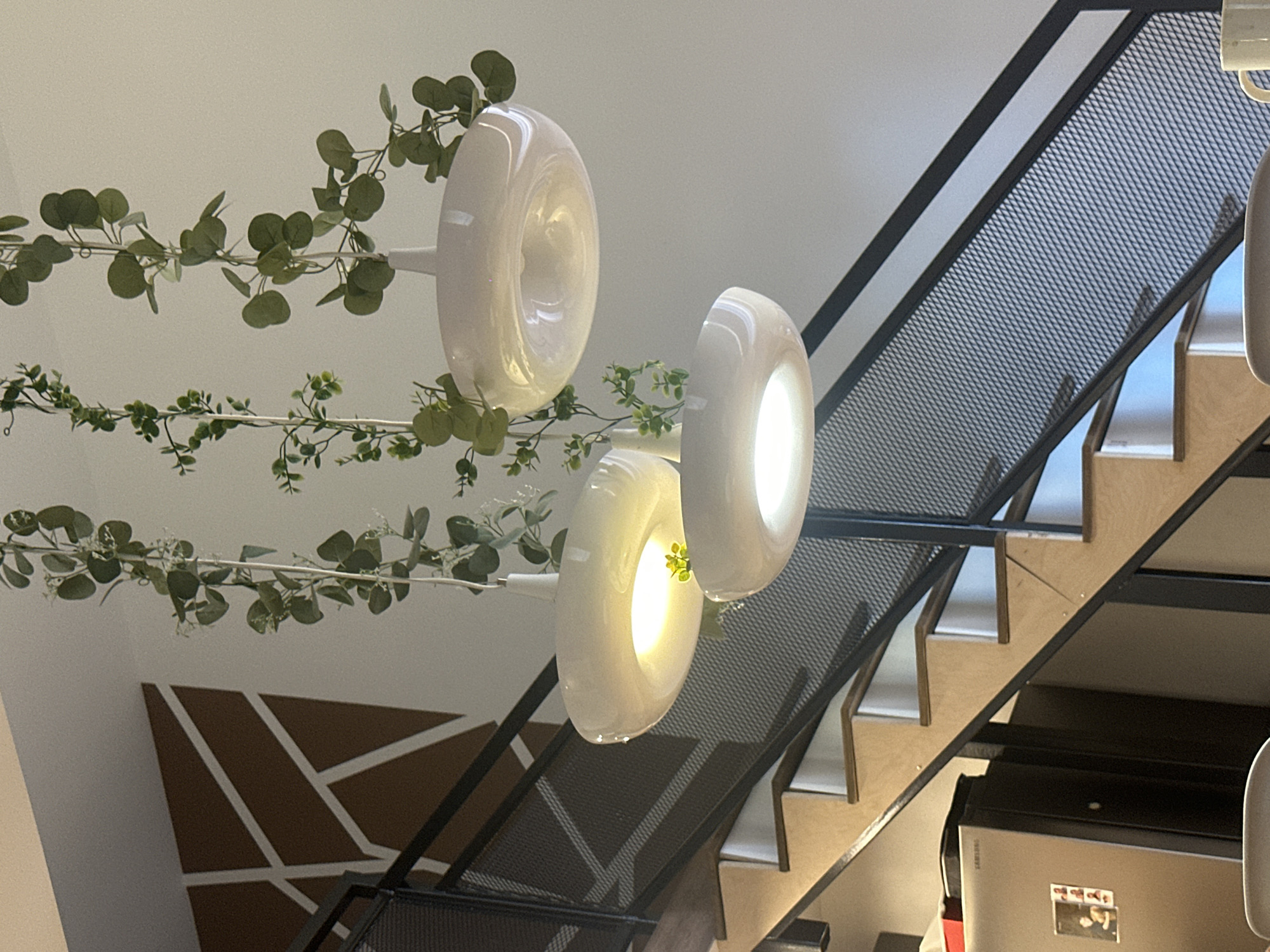 The image shows an interior view of a staircase with a modern design. The staircase features wooden steps and a black metal railing with a mesh-like pattern. Above the staircase, three modern pendant lights with white shades are suspended from the ceiling. Artificial greenery vines are draped around the lights and hanging down, adding a touch of nature to the decor. In the background, on the wall near the staircase, there is a geometric pattern in brown and white. To the left of the staircase, a silver refrigerator is visible. The overall scene suggests a contemporary living space. The location is unknown, Unknown.