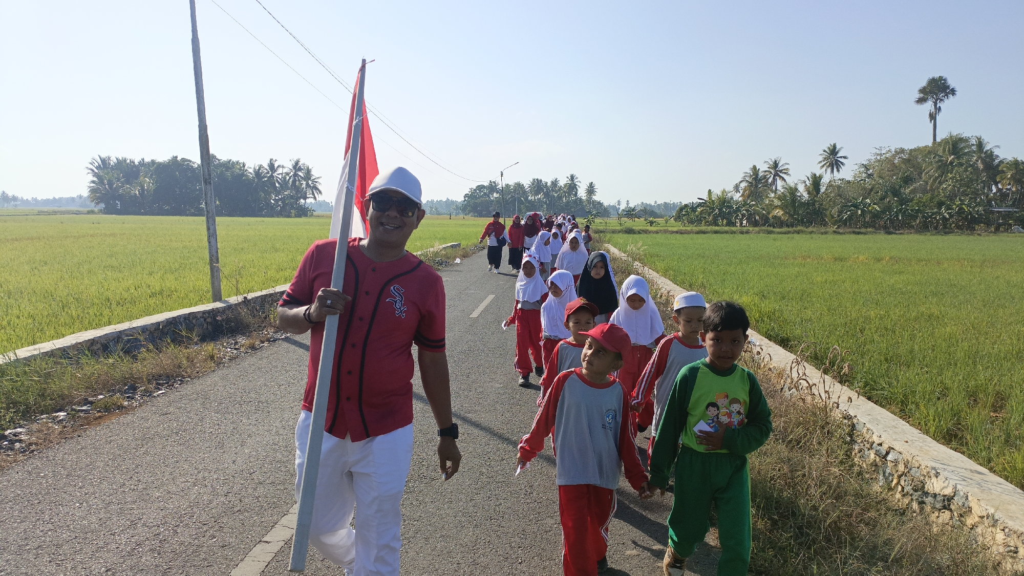 In the city of Batarang, Indonesia, a group of children and an adult are walking on a paved road. The adult, in the foreground, is wearing a red baseball jersey, white pants, sunglasses, and a white cap. He is holding a white pole with a red and white Indonesian flag. The children, following him, are a mix of boys and girls, some wearing white headscarves and red and white uniforms, others in green tracksuits. They are walking in a single file line. The road is bordered by a low stone wall and then stretches of green rice fields. Palm trees and other vegetation are visible in the background, under a clear blue sky. The scene appears to be a procession or a school outing in a rural setting.