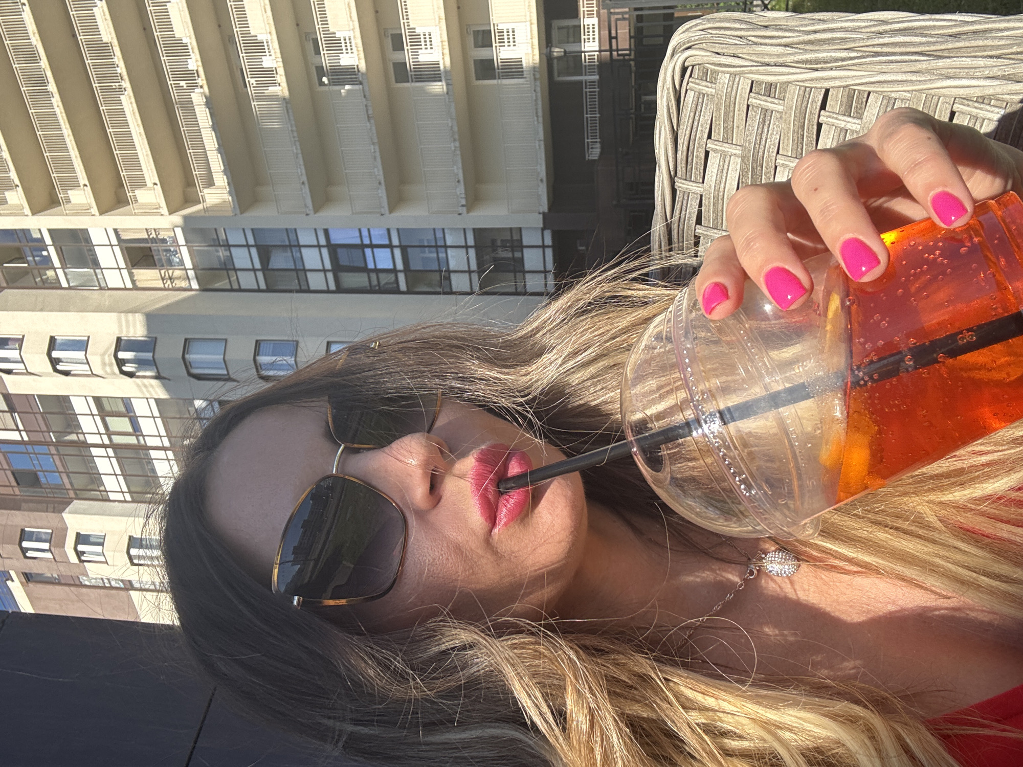 A woman is captured in a close-up, low-angle selfie, holding an orange-colored beverage in a clear plastic cup with a black straw. Her hand, adorned with bright pink nail polish, is visible near the top of the cup. She wears sunglasses with dark lenses and a thin gold frame, and her lips are painted a vibrant pink. A delicate silver necklace with a charm rests on her collarbone. Her blonde hair, with lighter streaks, frames her face and cascades around her shoulders.
In the background, a modern building with repeating balconies and windows is visible, suggesting an urban environment. The sunlight indicates it is daytime, likely sunny given the strong shadows. The scene appears to be outdoors, possibly on a balcony or patio, as a woven chair is partially visible. The overall impression is one of relaxation and enjoyment on a warm day. No visible text is present in the image. The context suggests the location is Saint Petersburg, Russia.