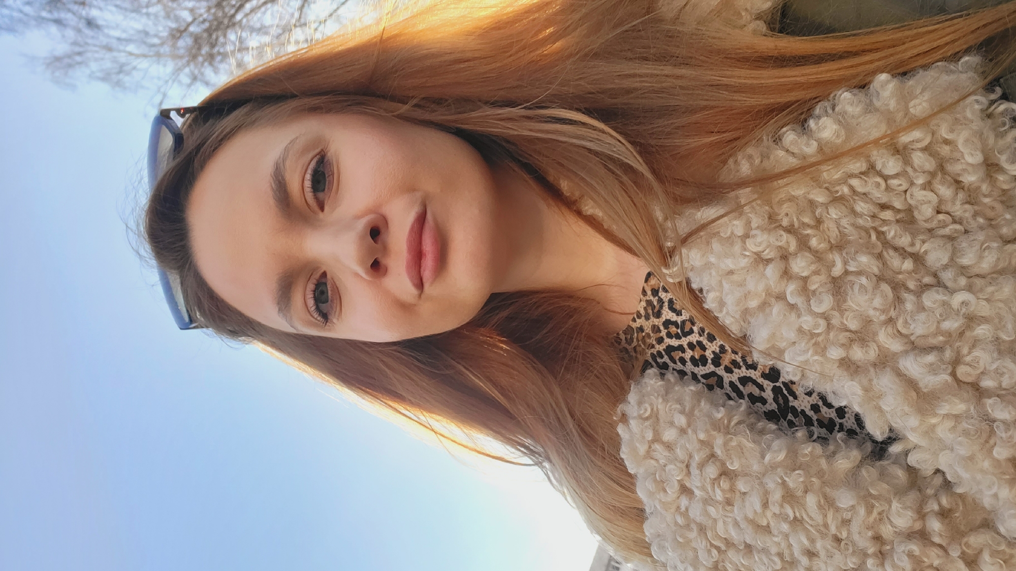 A young woman is pictured in a selfie. She has long, reddish-blonde hair. She is wearing sunglasses perched on her head and a sheepskin or faux-sheepskin jacket over a leopard-print top. The background is a clear blue sky and the blurred branches of a tree. This appears to be an outdoor shot taken in daylight, likely during the daytime. The location in the city of Chaikovsky, Russia, is not apparent in this image.