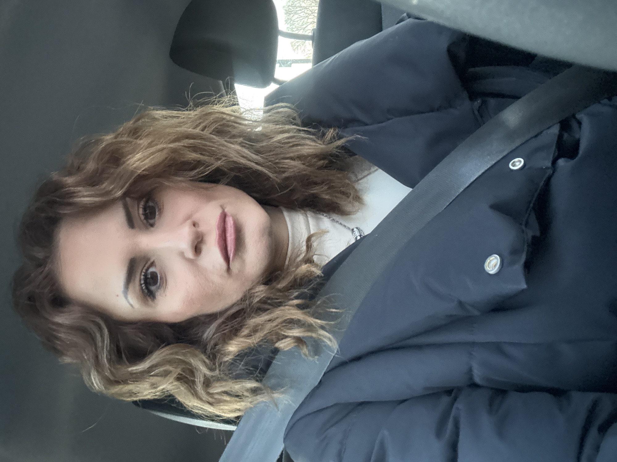 A woman with curly brown hair is wearing a navy blue puffer jacket and a seatbelt in a car. She is looking at the camera. The scene is likely indoors, in a vehicle. The location is unknown and there is no basis to conclude it is in London.