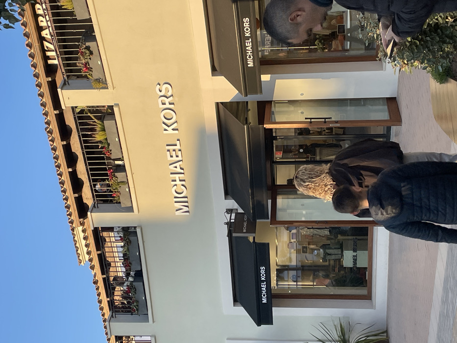 The image displays an outdoor scene at the Mallorca Fashion Outlet in Marratxí, Spain. The primary subject is the storefront of a Michael Kors store, identifiable by the large white "MICHAEL KORS" lettering on its light cream-colored facade. The store features multiple black awnings over its display windows and a double glass door entrance with brown wooden frames, which is open. Merchandise is visible through the store's glass front.
Above the Michael Kors store, a second story features a balcony with black wrought-iron railings and grey planter boxes containing green foliage and red flowers. A partial sign for "LIZARD" is visible on the upper level, to the right, behind the balcony railing. Several individuals are present on this balcony.
In the foreground, an adolescent male with short brown hair, wearing a dark blue puffer jacket, stands with his back to the camera. Immediately in front of him, a female with curly blonde hair, wearing a dark jacket, is also seen from behind, positioned near the entrance of the Michael Kors store. To the right, partially obscured by low green shrubbery, a male with short dark hair is visible in profile, looking down at a smartphone held in his hands. The ground is a light-colored paved walkway. The scene is illuminated by natural sunlight, casting soft shadows, and a clear blue sky is visible at the top.