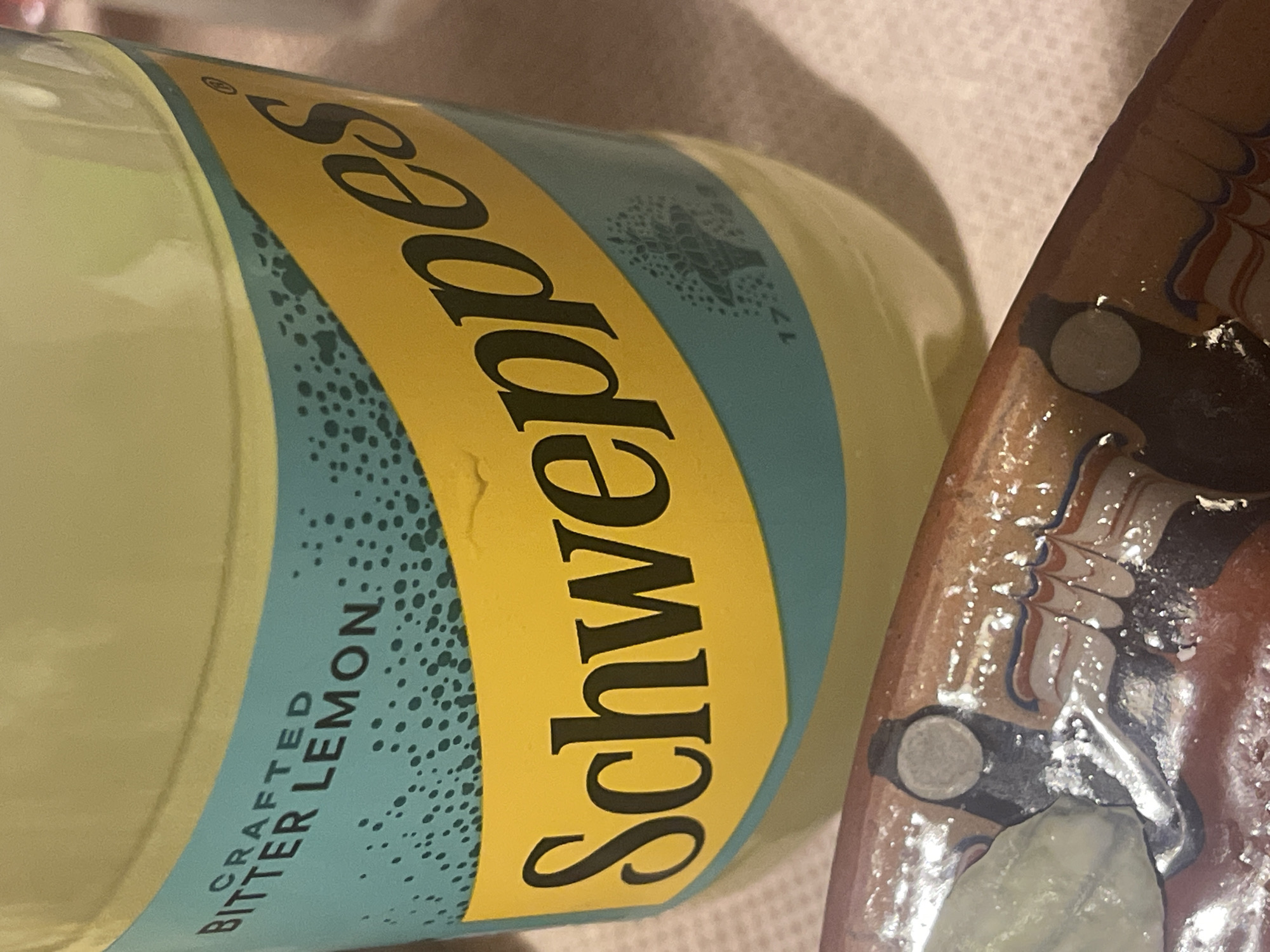 The image presents a close-up, angled view of a bottle of beverage and a portion of a decorative object, likely situated on a flat surface indoors.
The primary subject is a clear glass bottle containing a pale yellow liquid, identified by its label. The label features a prominent yellow section with the brand name "Schweppes" written in a distinctive, ornate black script. Adjacent to this, on a blue-green background adorned with numerous small, darker dots, the words "CRAFTED" and "BITTER LEMON" are printed vertically in a sans-serif black font. A registered trademark symbol (®) is visible near the "S" in "Schweppes". Further along the blue-green part of the label, a faint, intricate crest or tree-like emblem is visible, alongside the partial inscription "17__", suggesting a historical date.
To the right of the bottle, part of another object is visible. This appears to be a ceramic or pottery item, possibly a tray or plate, characterized by a glossy, reddish-brown base color. It is decorated with swirling patterns in white, blue, and reddish-orange hues. Several small, circular, silver-colored elements, resembling decorative studs or inlays, are incorporated into its design.
The background is softly blurred, appearing as a light-colored, possibly textured surface. The lighting is bright, illuminating the subjects and casting subtle shadows. No people or specific activities are depicted; the scene is static.