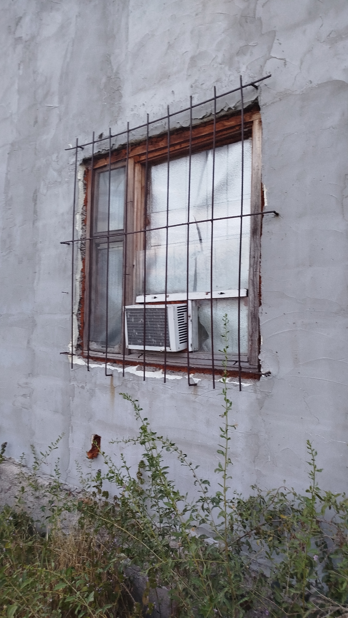 This is a medium shot, eye-level, outdoor, daylight, full-frame image of a window on a weathered, light gray, stucco building in Kostiantynivka, Ukraine. The window is secured with a dark metal grate, composed of vertical bars and horizontal crossbars. Inside the window, a white window air conditioning unit is visible, partially obscured by the grate. The window itself has wooden trim and panes that appear frosted or textured. Below the window, there is an area with overgrown dry grass and green leafy plants. To the left of the window, a small, red-brown brick opening is visible in the stucco wall. The overall condition of the building and its surroundings suggests neglect or disrepair.