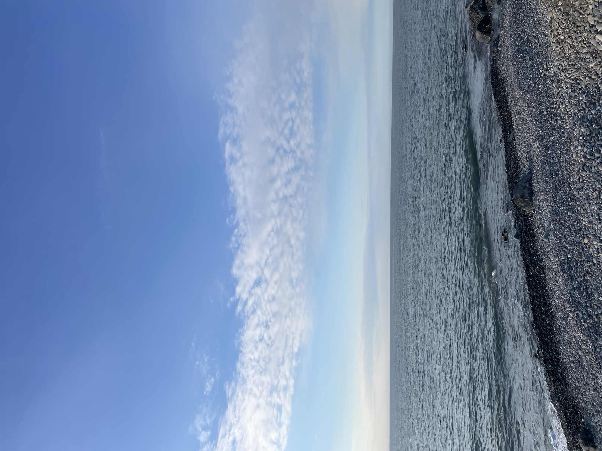 The image shows a coastline, likely a beach, in Batumi, Georgia. On the right side, the shore is covered with dark, small stones and pebbles. The Black Sea is to the left, with gentle waves lapping the shore. The sky takes up about half of the frame, showing a clear blue sky with scattered white clouds. No people or other distinctive objects are visible.