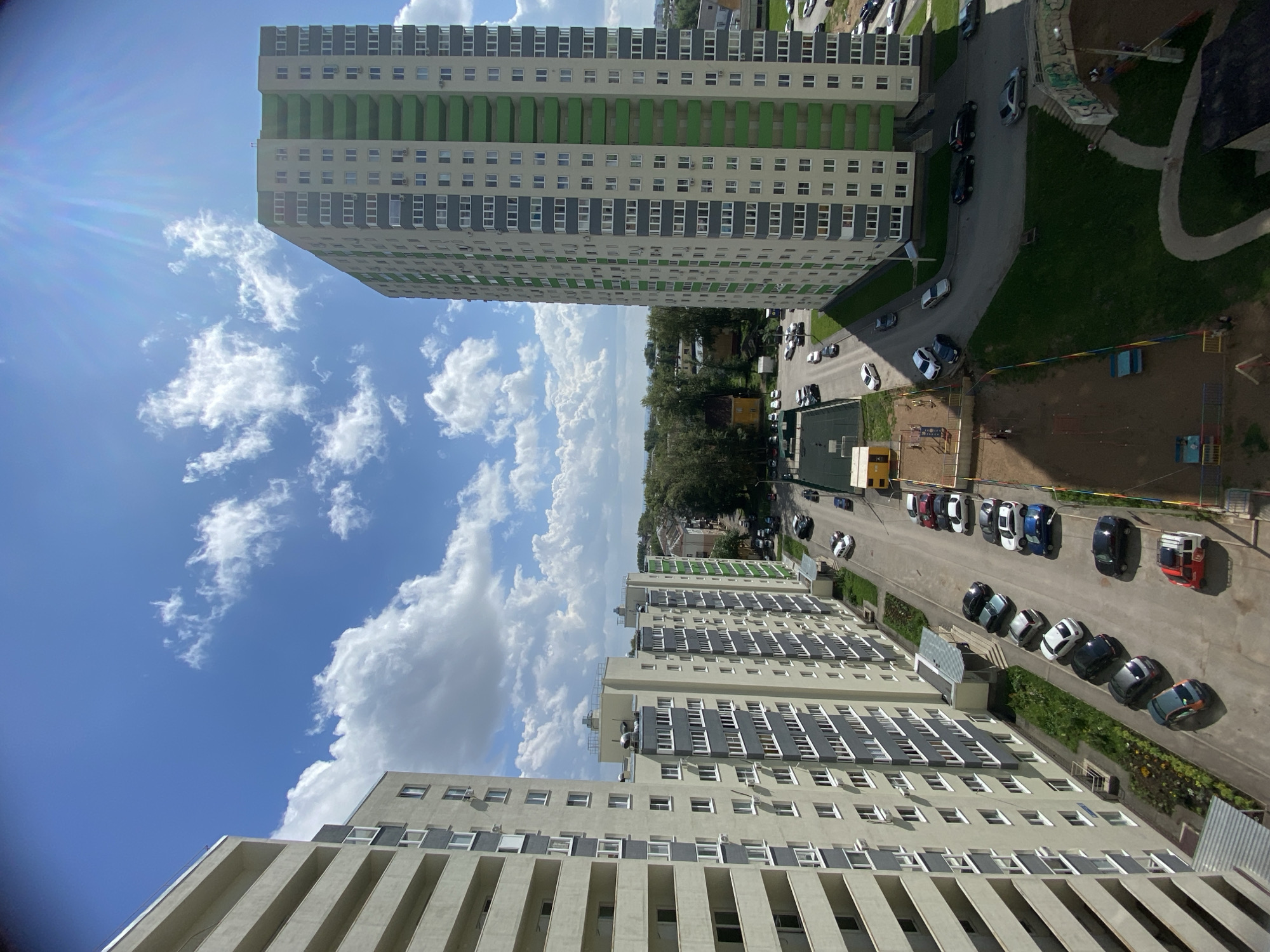 The image depicts a modern residential area in Ufa, Russia, on a sunny day with scattered clouds. Two prominent high-rise apartment buildings, one with light beige and grey facades and green balconies, and another with light beige and grey facades, dominate the scene. A street runs between the buildings, lined with parked cars. A playground with sand and colorful equipment is visible in the foreground. A basketball court is also present. Trees and other buildings are visible in the background, suggesting a well-established urban environment. The overall impression is one of a typical urban residential neighborhood.