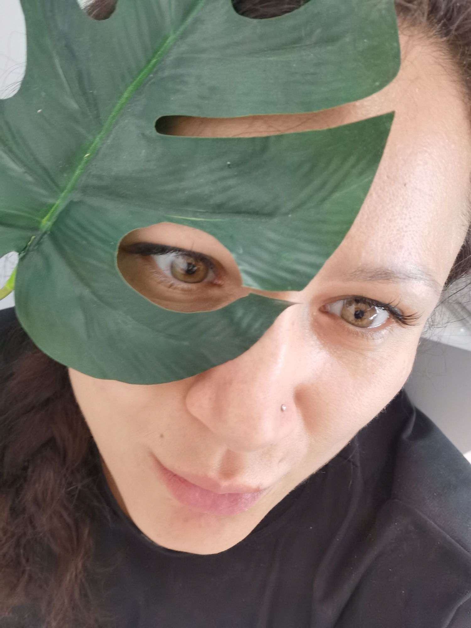 A woman is holding a large, green monstera leaf in front of her face. The leaf partially obscures her face, with holes in the leaf positioned to reveal her eyes. She has brown hair and a nose piercing. She is wearing a black t-shirt. The background is plain and white. There is no information to indicate that the image was taken in Chaikovsky, Russia.