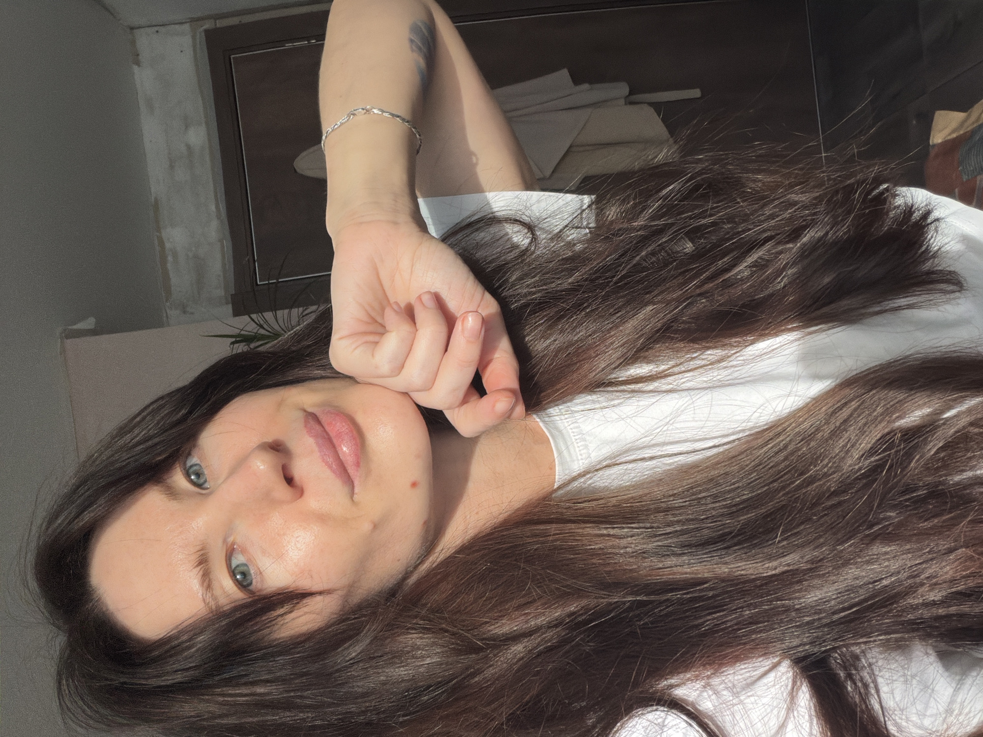 An adult female is depicted in a close-up, eye-level selfie taken indoors. She has long, dark brown hair, blue eyes, and a fair complexion with minor skin blemishes on her chin and right cheek. She wears a white crew-neck t-shirt. A silver-colored twisted chain bracelet is present on her left wrist, and a faded, dark-colored tattoo is visible on her upper left arm. Her left hand, with fingers curled into a loose fist, supports her chin. She looks directly at the camera with a subtle, closed-mouth smile. The background consists of an interior room with a light-colored wall exhibiting a horizontal band of unfinished or exposed material. A dark brown door and door frame are visible on the right side of the frame. A green houseplant is partially visible in the upper left background. Strong natural light illuminates the scene from the right, casting shadows. The specific location within Perm, Russia, is not identifiable from visual evidence.
