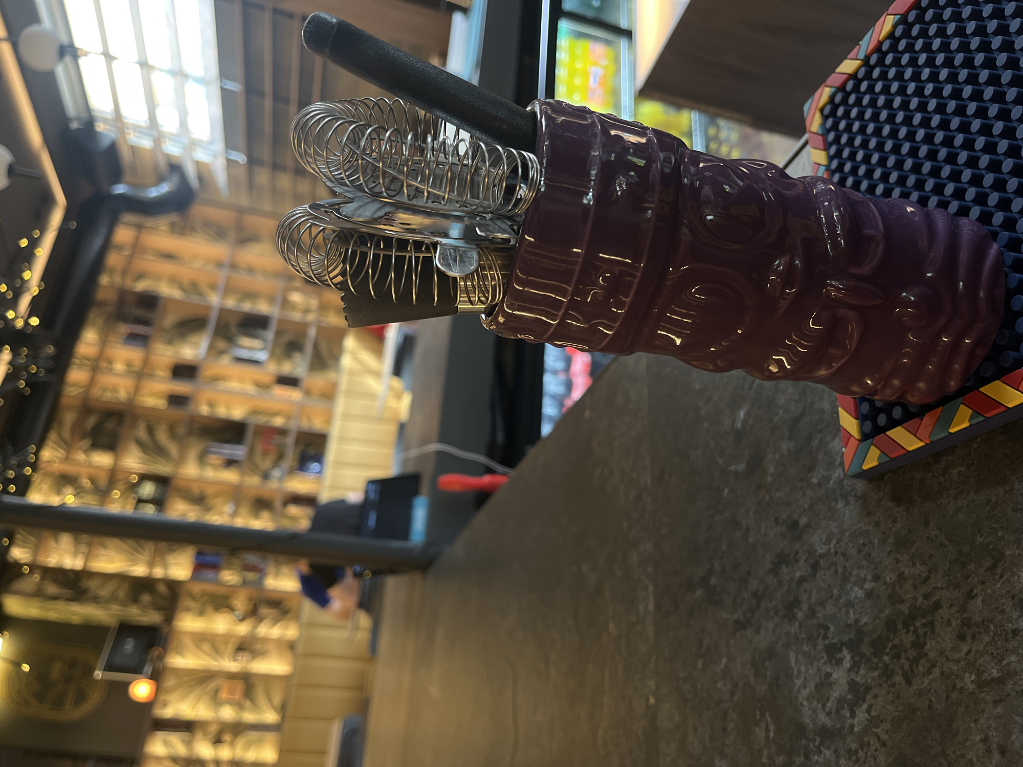 A close-up, low-angle shot focuses on a purple ceramic tiki mug filled with bar tools, including cocktail strainers and a black handled jigger, placed on a dark, speckled bar top. Behind the tiki mug, a blurred background reveals a dimly lit bar interior with golden, patterned wallpaper and shelves stocked with bottles. Two individuals are partially visible in the background, one wearing a blue shirt and the other a black shirt, both seated at the bar. The overall impression is a dimly lit, atmospheric bar.