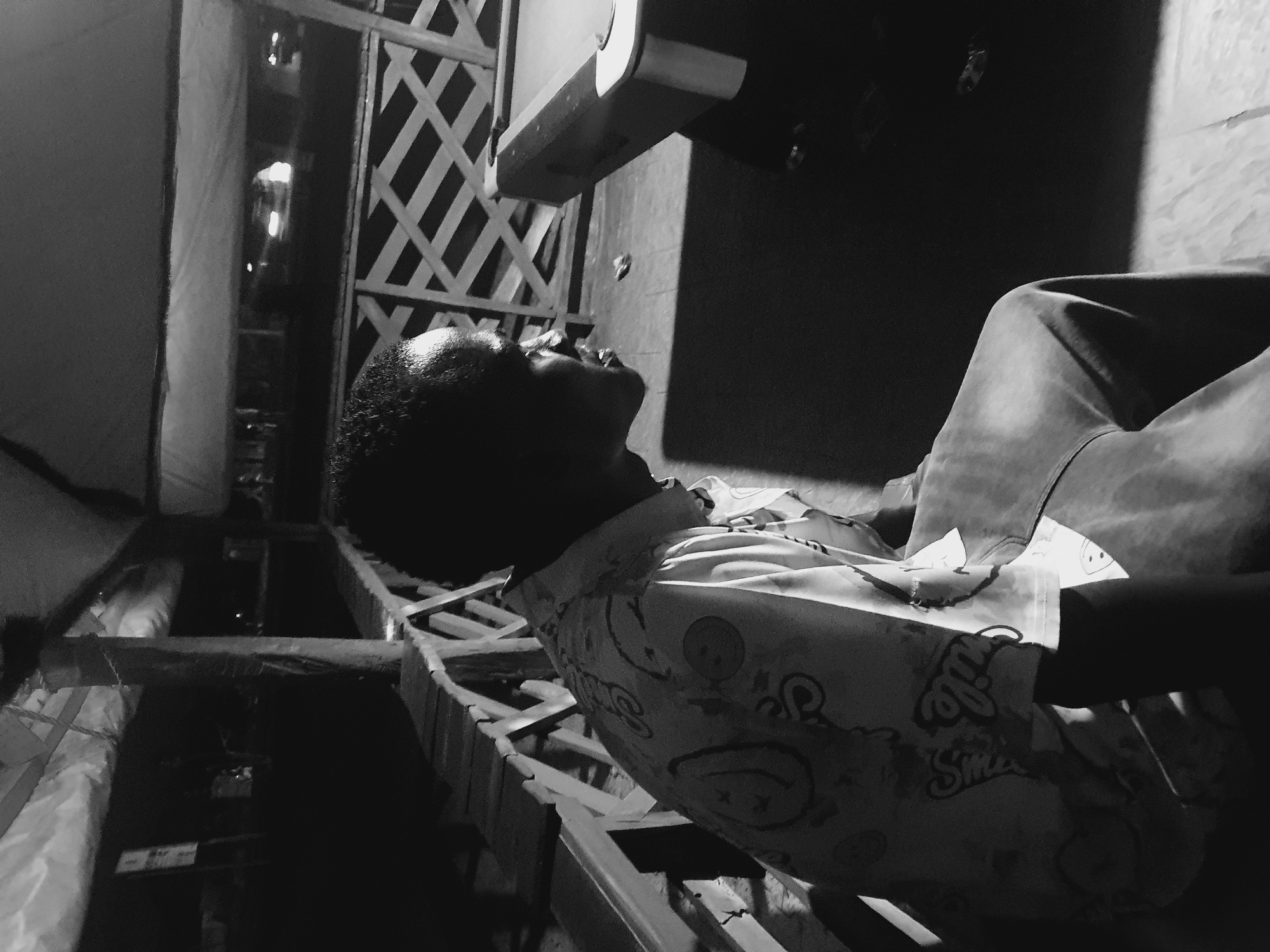 A black and white low-angle photograph depicts a person reclining on a wooden structure resembling a porch or deck. The person is wearing light-colored patterned clothing. The head is tilted upwards. A dark rectangular object is positioned above the individual. The surrounding environment includes a latticework barrier. Based on the location, the scene could be a residential or recreational space in Lokoja, Nigeria.