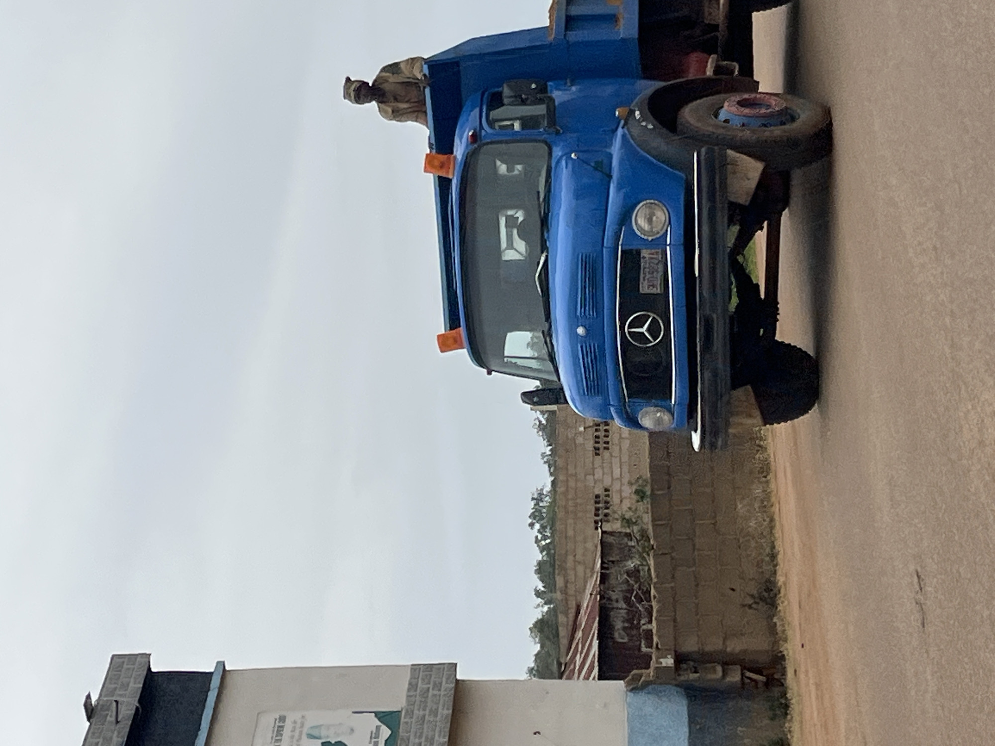 In Bukuru, Nigeria, a blue dump truck is parked on a dusty road. A person wearing a brown jacket and a head covering sits atop the truck's cab, looking out. The truck has an orange flashing light on its roof and a visible Mercedes-Benz logo on its grille. A building with a light blue and white facade is on the left side of the image, with a poster visible on its exterior. Behind the truck, a brick wall and a corrugated metal roof structure are visible, suggesting a residential or industrial area. The sky is overcast.