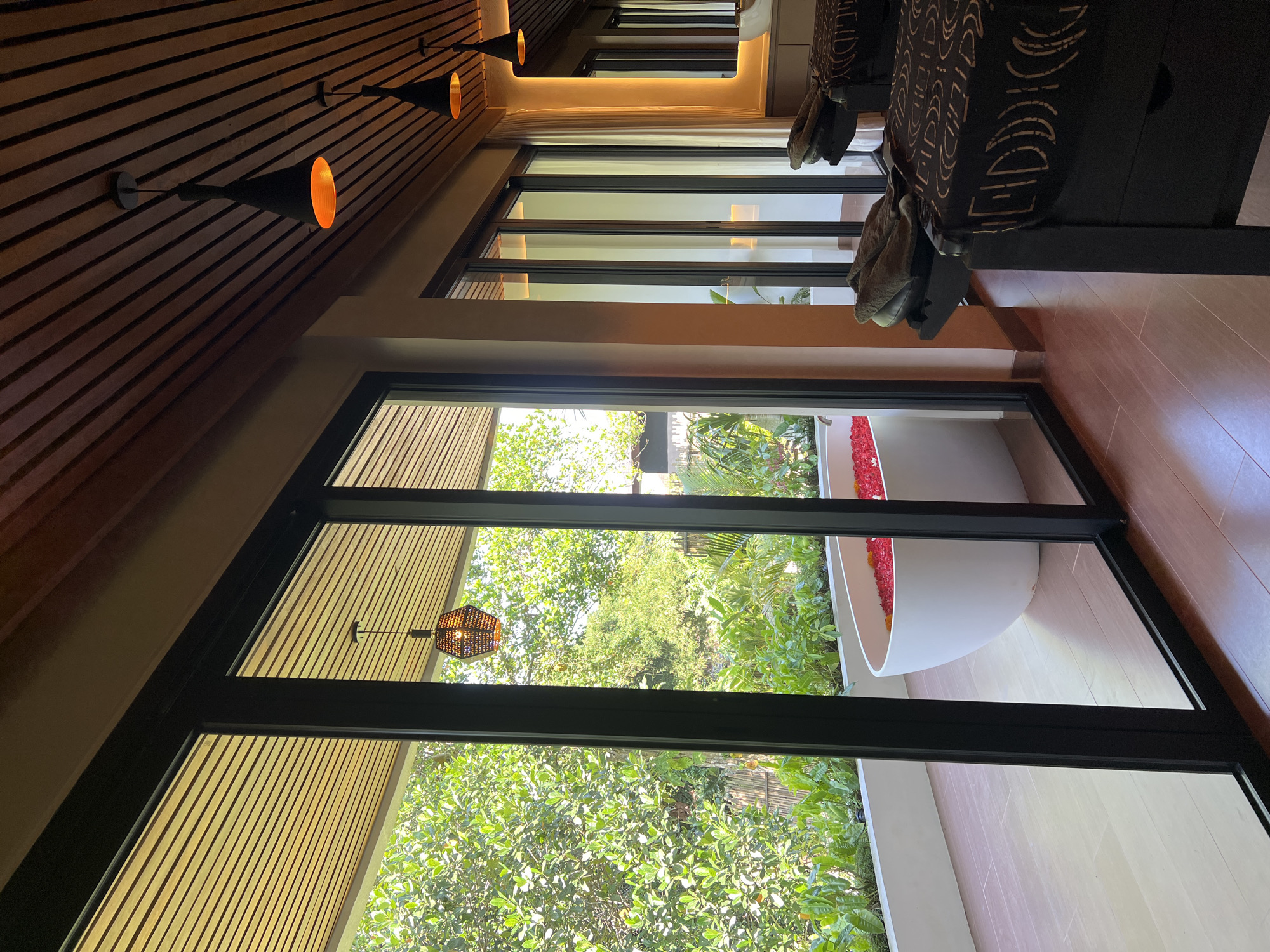 In a spa in Ubud District, Indonesia, a serene scene unfolds. Through a large glass sliding door, a white bathtub filled with red flower petals and yellow flowers sits on a wooden deck surrounded by lush greenery. Inside the spa, two massage beds with dark patterned coverings and folded towels are visible. The room features a wooden slatted ceiling with hanging pendant lights, casting a warm glow. The overall ambiance suggests a tranquil and luxurious spa experience, possibly a private suite or treatment room with an outdoor bathing area.