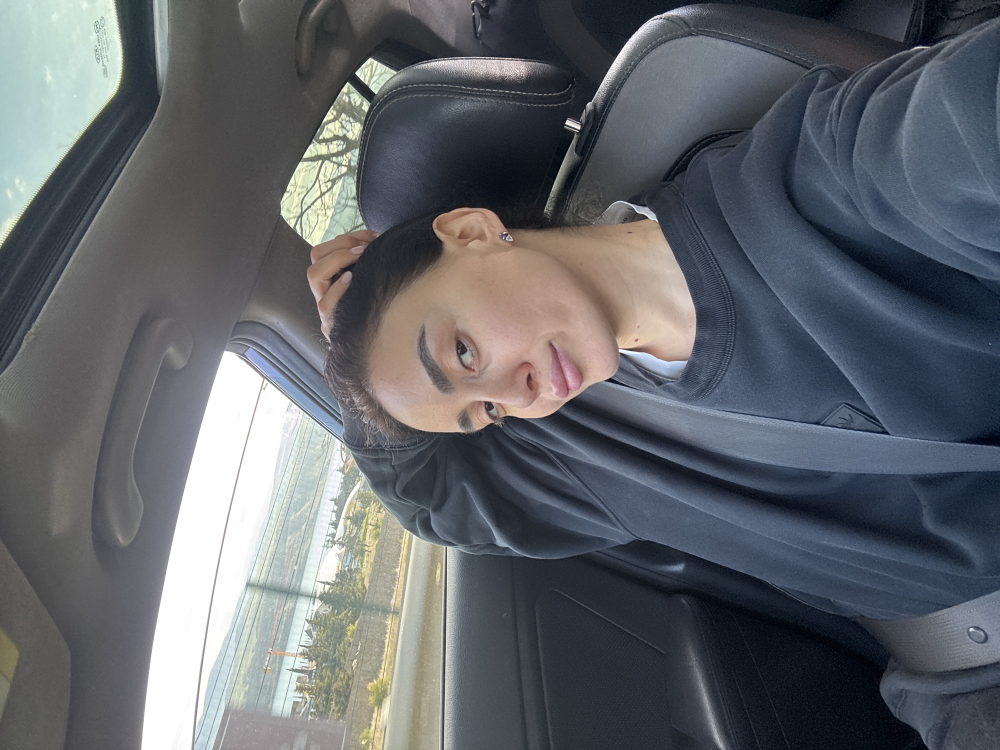 A woman is seated in the passenger seat of a car, wearing a gray sweatshirt and seatbelt. The background shows a view of Tbilisi, Georgia, through the car window. Trees and buildings are visible in the cityscape. The car appears to be in motion. The woman has dark hair, her right hand is above her right ear, and a stud earring is visible on her right ear.