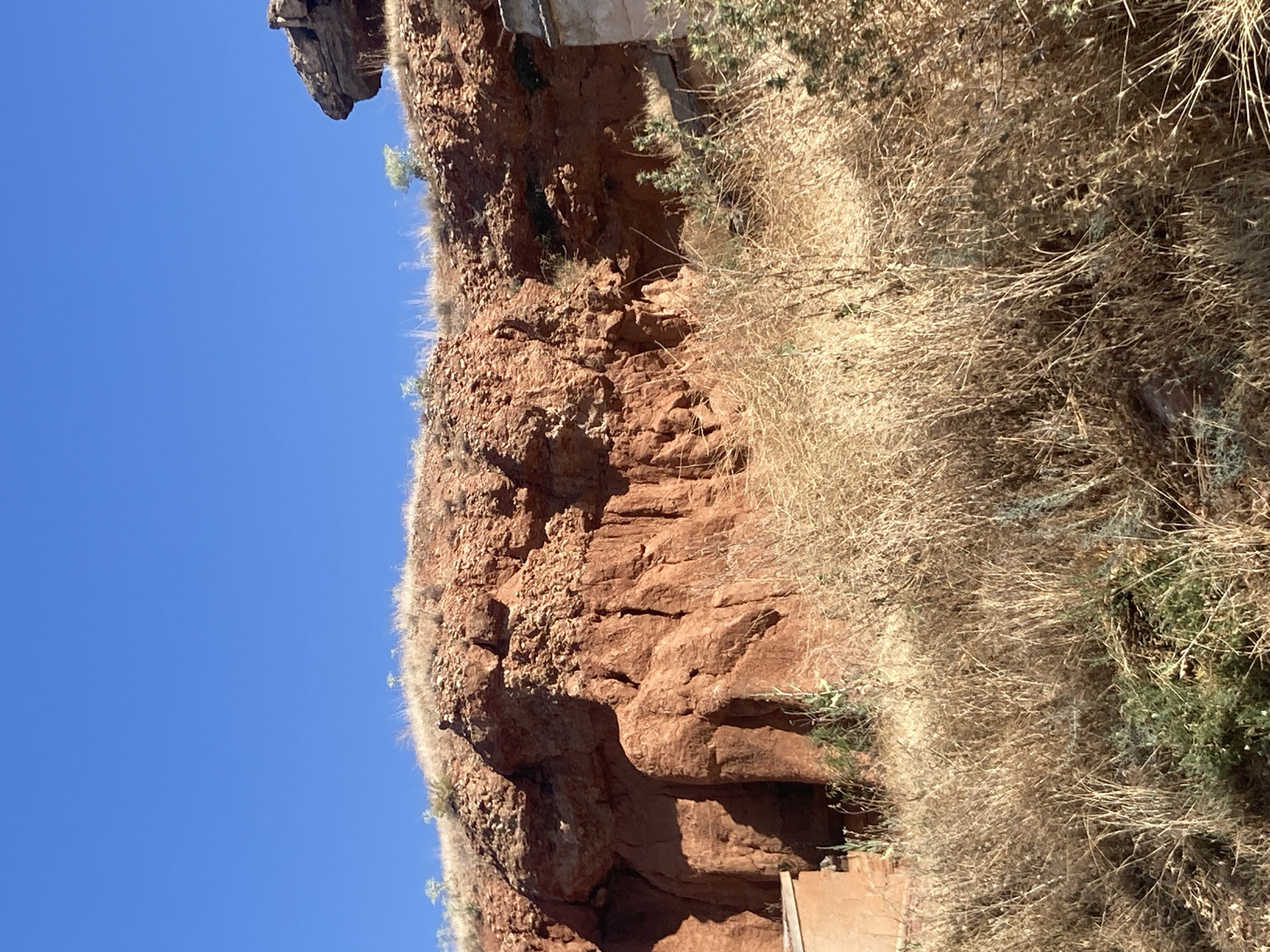 The image depicts a natural landscape with a prominent geological formation of reddish-brown rock, likely sandstone or clay, in Avanos, Turkey. The rock face is uneven and weathered, showing signs of erosion with visible layers and crevices. Vegetation, consisting of dry grasses and some sparse green bushes, covers the foreground and the slopes leading up to the rock. The sky is a clear, bright blue. On the left side of the frame, a portion of a man-made structure, possibly a building with a tan-colored wall, is visible at the base of the rock formation. On the right side, another part of a building, appearing to be made of stone or a similar material with a lighter facade, is partially visible. The overall scene suggests a dry, arid environment typical of the Cappadocia region.