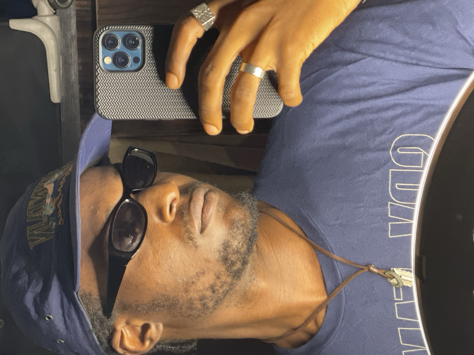 A close-up, waist-up, selfie-style photograph shows a dark-skinned male wearing a blue baseball cap, black sunglasses, and a navy blue t-shirt. The man has a goatee and a thin mustache. He is holding a smartphone in his right hand, which is angled towards the camera. The phone case has a chevron pattern. He is wearing two silver rings on his right hand; one on the ring finger and another on the middle finger. A thin brown leather cord necklace with a metallic pendant is visible around his neck. The t-shirt has a graphic design on it consisting of large, stylized letters. The background is dark and appears to be indoors, possibly a room or closet, with shelving and an object that looks like a white plastic brace or shelf support visible on the top right. The image is dimly lit, with a warm light source that appears to be coming from below and slightly to the left, casting shadows on the man's face and neck. There is no discernible indication of the location being in Abuja, Nigeria.