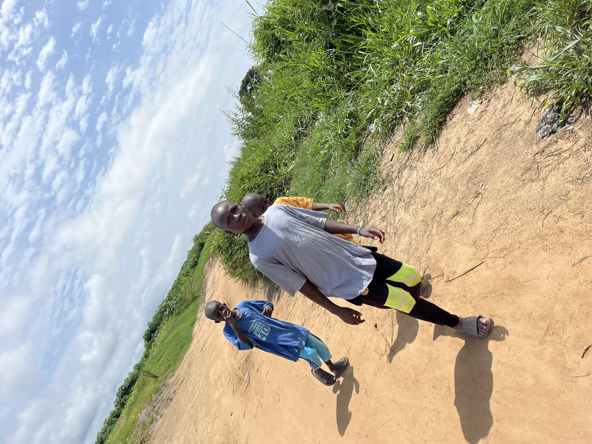 Three young males are captured standing on a reddish-brown dirt path in an outdoor, sunny setting, likely in Idah, Nigeria. The image is rotated clockwise, with the horizon appearing diagonal.
In the foreground and center, a boy with short, dark hair wears a light gray t-shirt, black pants with bright yellow-green panels, and gray sandals, looking directly at the camera with a neutral expression. Behind him and slightly to the right, another boy is partially visible in a yellow t-shirt. Further back on the left, a third, smaller boy wears a blue t-shirt with "YOUTHPAD DOUBLE MINT" and "40" printed on it, light blue-green pants, and dark sandals; he also faces the camera, possibly with a slight smile.
The boys are standing on a wide, unpaved path, likely a rural road or track. Lush, dense green vegetation, consisting of tall grasses and shrubs, lines both sides of the path, indicating a tropical environment. The sky in the upper left features scattered white clouds against a bright blue background. Strong shadows cast by the boys on the path suggest it is mid-morning to early afternoon. Small pieces of litter are visible on the dirt path near the vegetation. The scene depicts a casual moment, possibly during a walk.