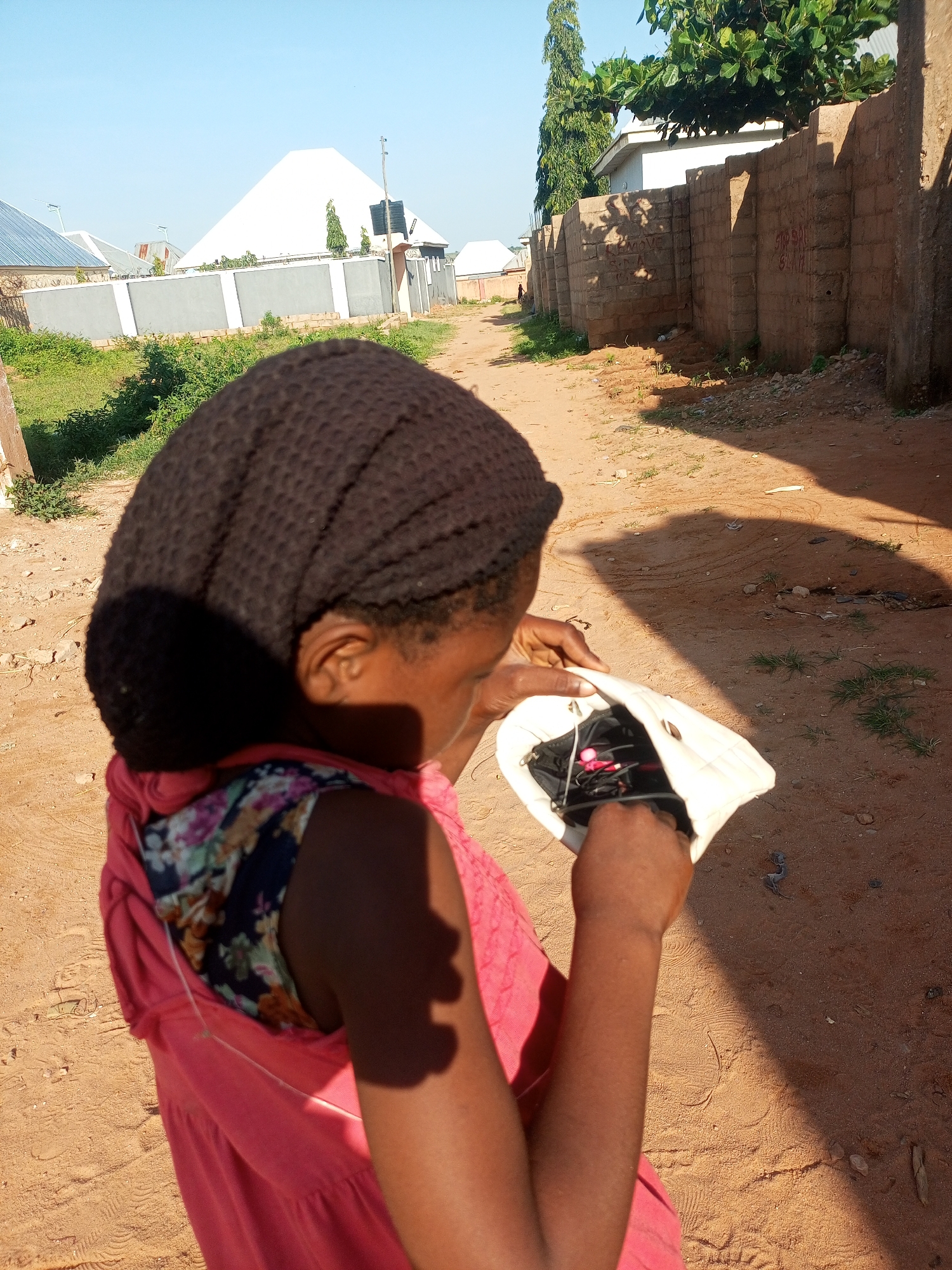 The image captures a person from behind, wearing a dark, textured headscarf and a pink dress with a floral pattern underneath. The individual is holding a small, light-colored bag open, with what appears to be a phone and earbuds inside. They are standing on a dirt path in what looks like a residential area with some buildings visible in the background. The environment suggests a sunny day, with a clear blue sky and shadows cast on the ground. The buildings in the distance include structures with pitched roofs and walls constructed from bricks and concrete. There is also greenery and vegetation present, including trees and bushes. The overall scene depicts an outdoor setting in a possibly rural or semi-urban part of Tempure, Nigeria.
