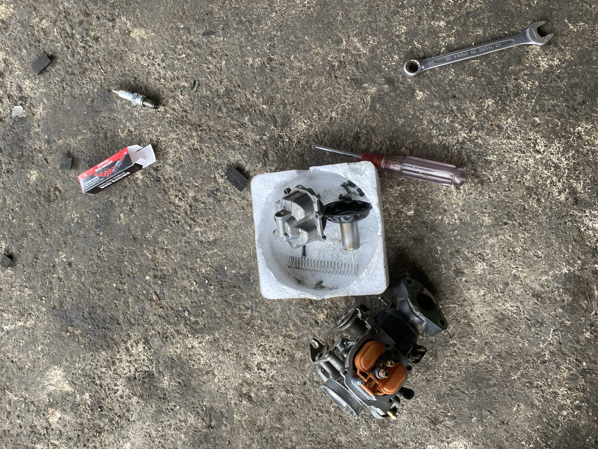This image shows a top-down view of a concrete surface with various motorcycle carburetor parts and tools scattered on it. In the upper right corner, a spark plug and its red and black packaging are visible. A disassembled carburetor, featuring orange float bowls, is positioned in the lower left. To its right, a white styrofoam container holds some carburetor components, including a spring and a metal rod. A screwdriver with a red handle and a silver wrench are also present on the concrete. The ground appears worn and somewhat dirty, suggesting an outdoor or workshop environment. There are no people visible in the image.