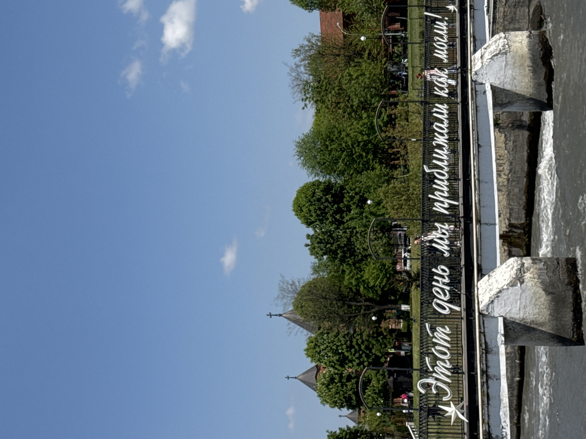 The image shows a scene in Vladikavkaz, Russia, near the Terek River. A low dam or weir stretches across the river, with water cascading over its edge. On the near side of the dam, a wrought-iron fence runs parallel to the water's edge. White lettering on the fence spells out a message in Russian. Behind the fence, trees and buildings are visible. People are present in the background, though they appear relatively small in the frame. The sky is blue with a few scattered clouds.