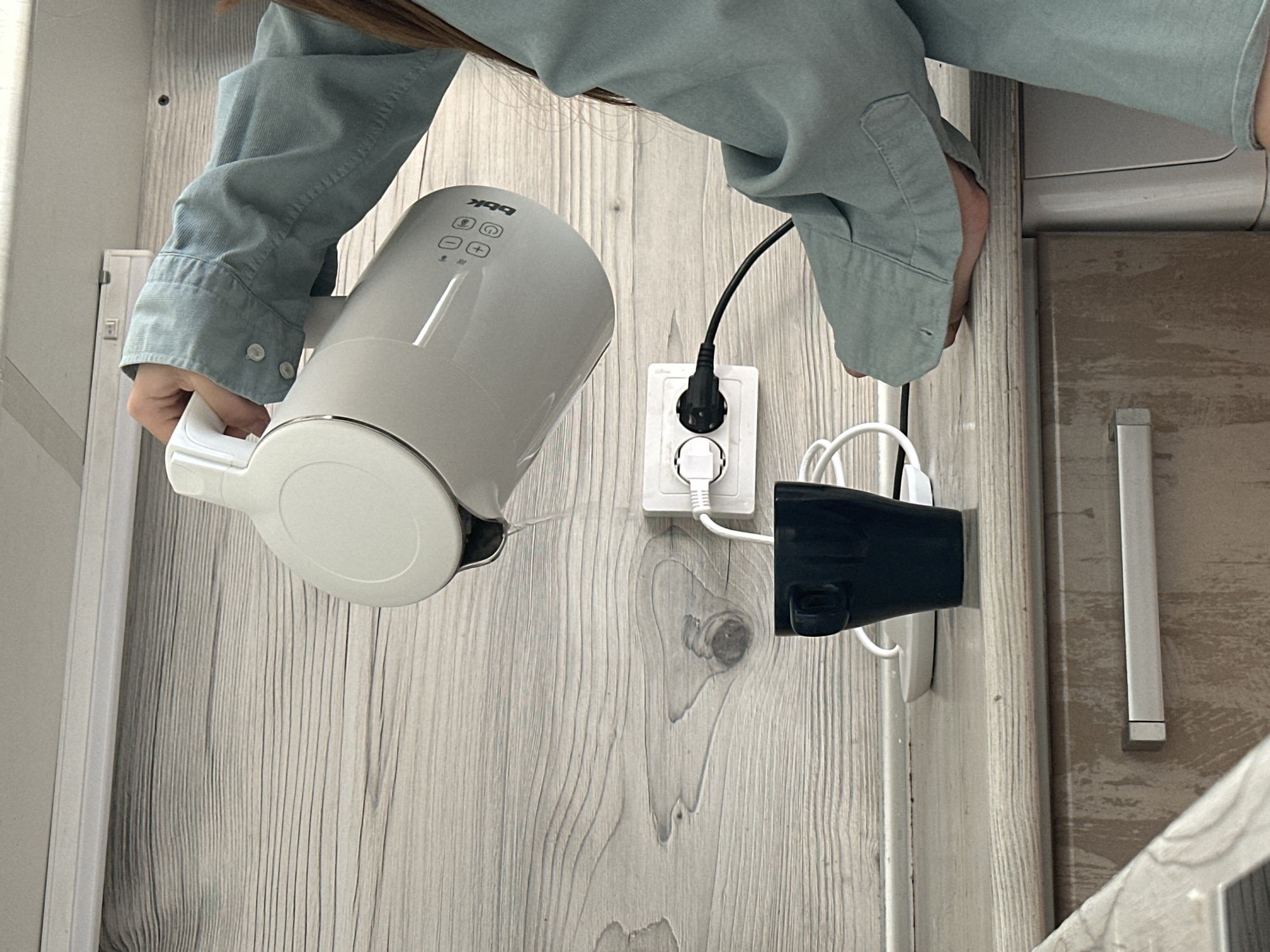 A person wearing a light blue shirt holds a white electric kettle. The kettle is positioned near a white electrical outlet, which has two plugs inserted. One plug is black and connected to what appears to be a black hair dryer. The scene is indoors with a light grey, wood-grained wall as the background. A portion of a door is visible on the right side of the image, featuring a silver handle. The location is Chaikovsky, Russia.