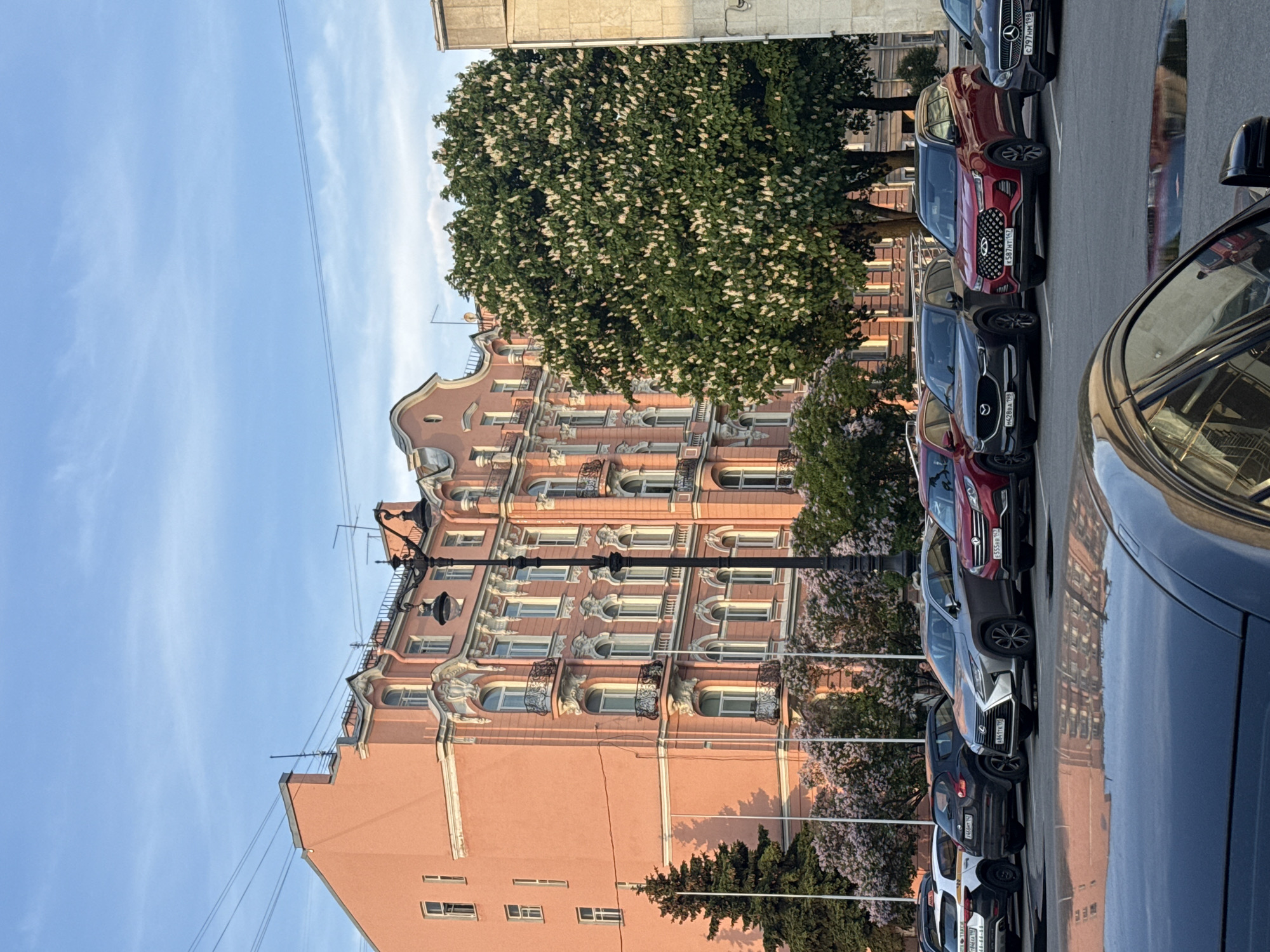 The image showcases a street scene in Saint Petersburg, Russia, characterized by a peach-colored residential building of architectural interest and a row of parked cars along the street. The building is the focal point, presenting ornate window designs and decorative details on its facade. Several trees, one with blossoming white flowers, add to the urban landscape. Parked cars, some of which are partially visible, line the roadside; their registration plates show Russian vehicle registrations. The sky is a clear blue with a few wispy clouds, indicating a fair weather day. The scene offers a glimpse of typical urban life in this historical city.