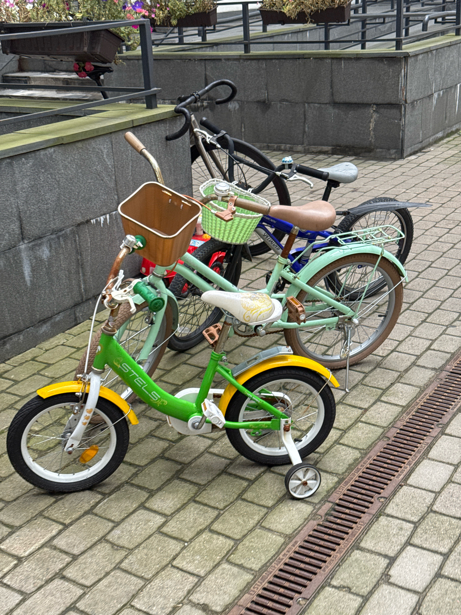 Two bicycles and a child's bicycle are parked on a brick sidewalk next to a concrete wall. The child's bicycle is green and yellow and has training wheels. The other two bicycles are darker in color and are larger. The scene appears to be in an urban environment, possibly a park or a residential area. There are no people visible in the image. The background shows some greenery and a metal railing. The lighting suggests it is daytime.