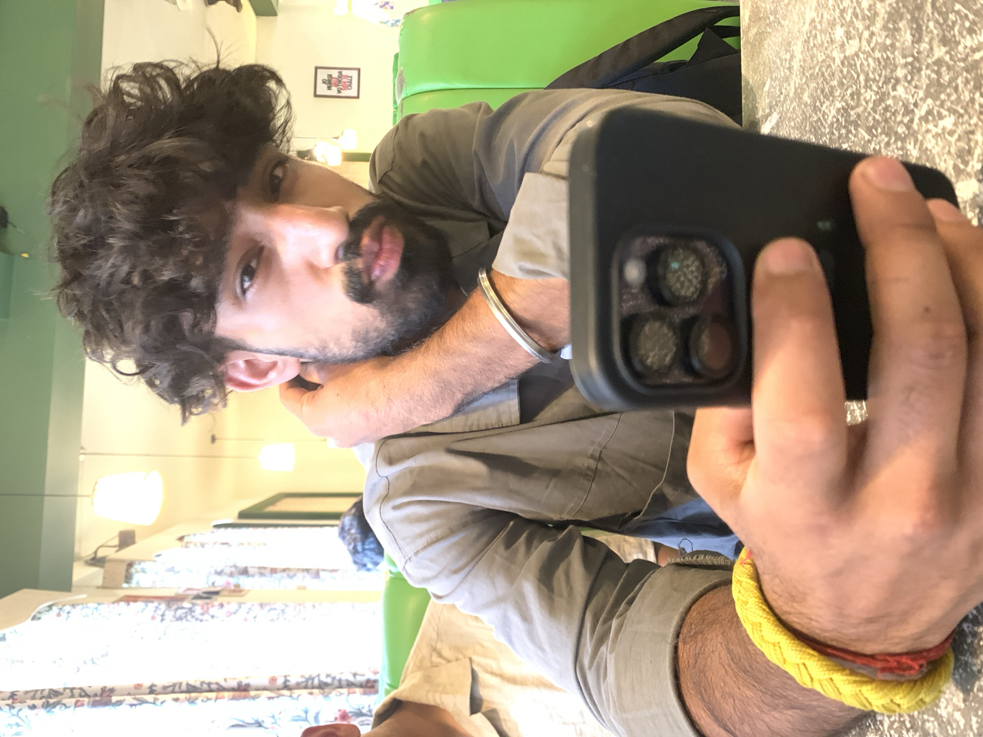 A man with curly dark hair, a beard, and a mustache is holding a smartphone in the foreground, looking directly at the camera. He is wearing a grey collared shirt and has a silver bracelet on his left wrist and a yellow and red braided bracelet on his right wrist. The background shows a brightly lit interior with green booth seating and patterned curtains. In the distance, a framed poster with text is visible on the wall. The overall setting suggests a cafe or restaurant.