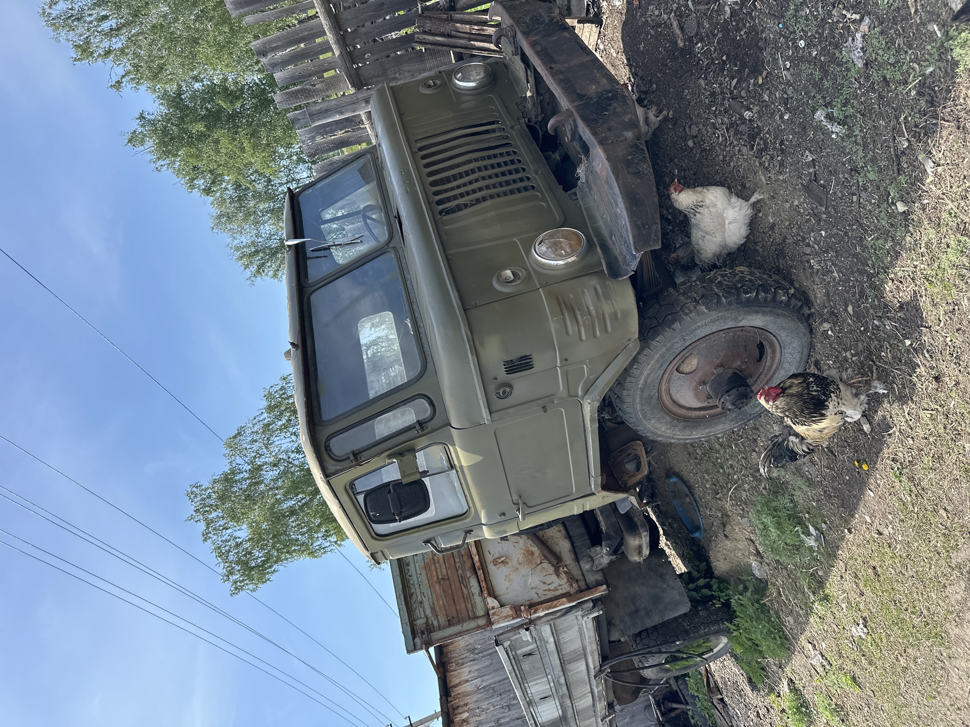 The image depicts an old, olive-green truck parked on a patch of dirt and grass in an unkempt rural setting. A weathered wooden fence and scattered trash are visible in the background. Two chickens, one white and one black and white, are foraging near the truck's tire. The sky is blue with a few trees visible in the background. Power lines are also noticeable in the background. The location is in the city of Unknown, Unknown.