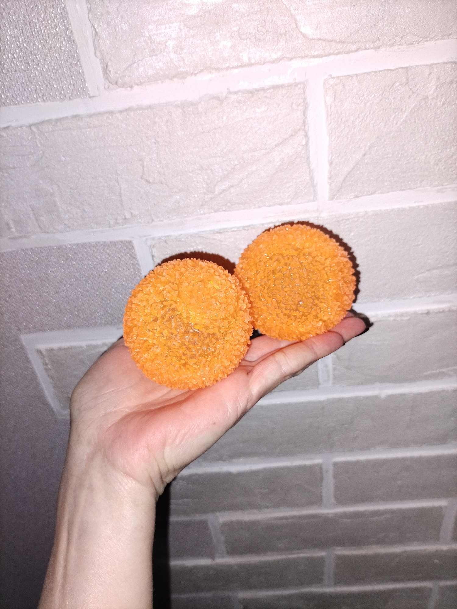 A human hand holds two bright orange, spherical objects against a textured wall background. The hand is positioned palm-up, cradling the two round items, which have a spiky or granular surface texture, resembling glitter or small crystal shards. The objects cast soft shadows on the hand and the wall behind. The person's fingernails on the visible fingers are painted black.
The setting appears to be indoors, with a wall featuring a light-colored, faux-brick pattern. The "bricks" are a mix of off-white and light gray, separated by white grout lines. In the upper left portion of the background, a section of the wall exhibits a different, more finely textured or perhaps slightly reflective pattern, distinct from the brick-like texture. The lighting is artificial, creating shadows and highlighting the texture of the objects and the wall. There is no visible text in the image.