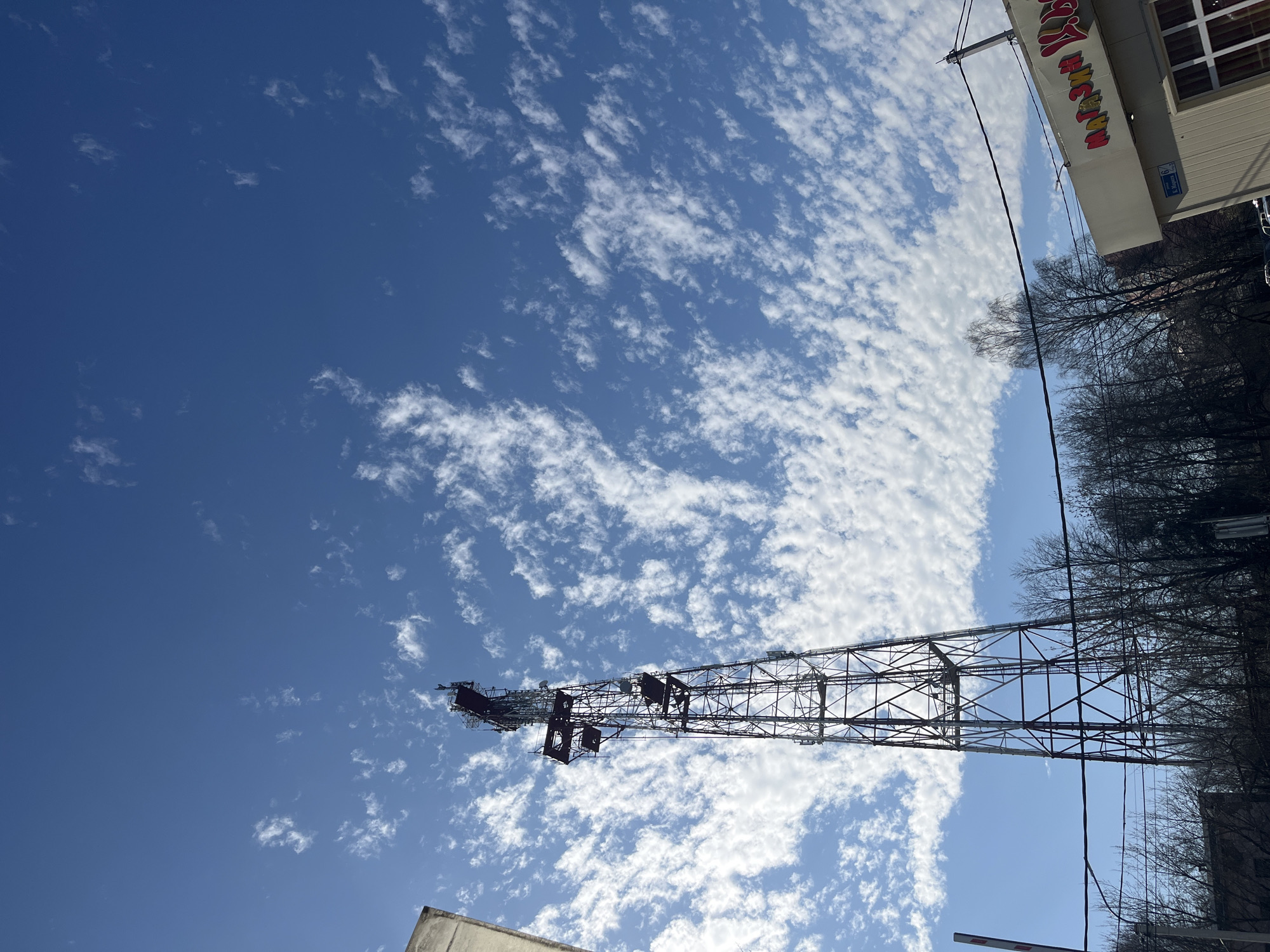 The image shows a communications tower against a blue sky with scattered white clouds. The tower is constructed of metal latticework and extends diagonally across the frame. Bare trees are visible to the right of the tower, and the corner of a building with a sign written in Russian is visible at the top right of the frame. The location is in the city of Klin, Russia.