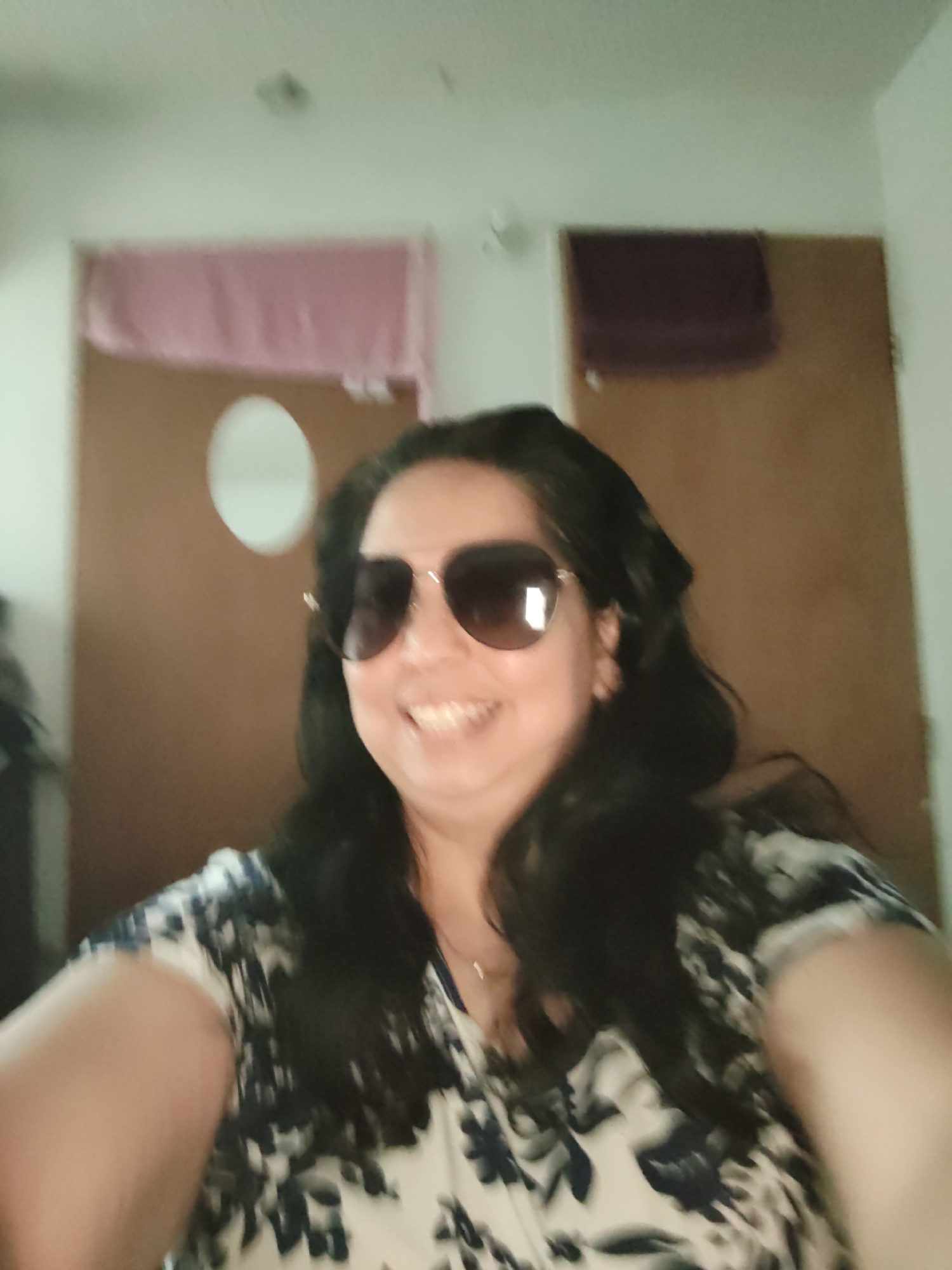 The image shows a woman wearing sunglasses and a patterned dress. She is smiling and appears to be taking a selfie. The background is out of focus and shows a wall with two framed items, one pink and one dark purple, and a circular object. The overall impression is a casual, indoor setting. There is no information in the image that allows for the identification of its location in Piloncito, Venezuela.