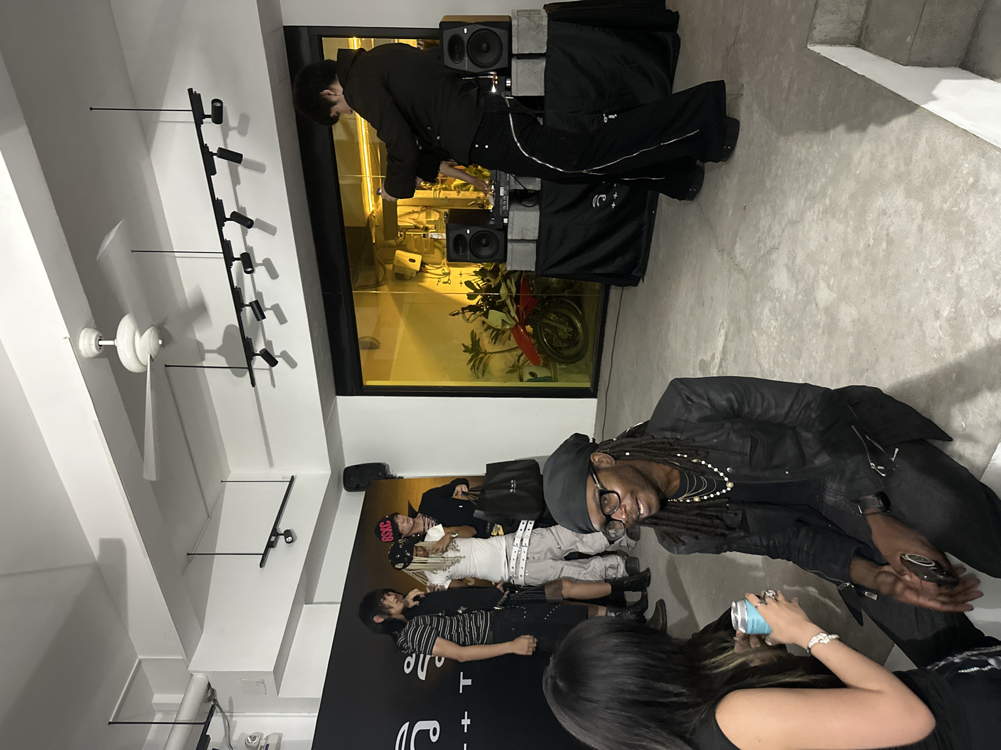 The image depicts an indoor event, likely a gathering or small party, possibly at a gallery or studio space. The setting appears to be modern, with exposed concrete floors and a white ceiling featuring track lighting and a ceiling fan. The background wall is adorned with a large black banner displaying white text and symbols, potentially indicating branding or an event title. To the right, a large window with yellow lighting illuminates an interior space, where a dirt bike is visible, suggesting a unique or artistic display.
In the foreground, two individuals are seated. One, on the left, has long dark hair and is wearing a black sleeveless top. They are holding a blue can. The other individual, in the center, is wearing glasses, a black jacket, and multiple necklaces, and is looking to their left, engaged in conversation.
In the middle ground, a group of four individuals are standing near the black banner. One person on the far left is wearing a striped t-shirt and dark pants. Next to them, two individuals are dressed in a more avant-garde fashion, with one in a white top and cargo pants, and another in a black top and hat. The fourth individual on the right is wearing a black outfit and holding a black bag.
Further to the right, another individual is standing behind a DJ setup, which includes turntables and speakers placed on concrete blocks. This person is dressed in black and appears to be interacting with the equipment. The overall atmosphere suggests a casual yet stylish event, possibly a launch party, exhibition opening, or a social gathering with a creative theme.
The image does not provide any explicit visual cues that would definitively place the event in Thủ Đức, Vietnam.