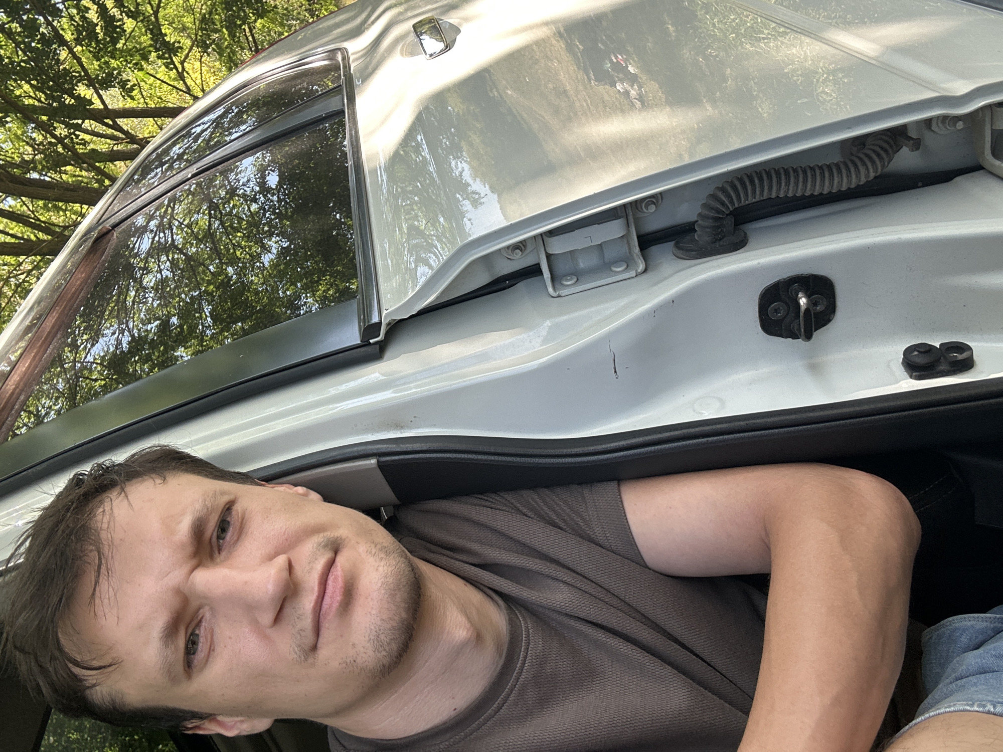 A close-up, eye-level selfie shows a young man with short, dark hair, light skin, and a neutral expression. He is looking directly at the camera, with his left arm resting on his lap. He is wearing a brown, textured t-shirt and denim shorts. He is sitting in the open driver's side door of a white car, which is parked outdoors with trees and foliage visible in the background, suggesting a natural, possibly rural, setting. The car door is slightly ajar, revealing the car's interior and exterior metalwork, including a black rubber hose and door latch mechanism. The lighting suggests it is daytime, with sunlight reflecting off the car's surface.