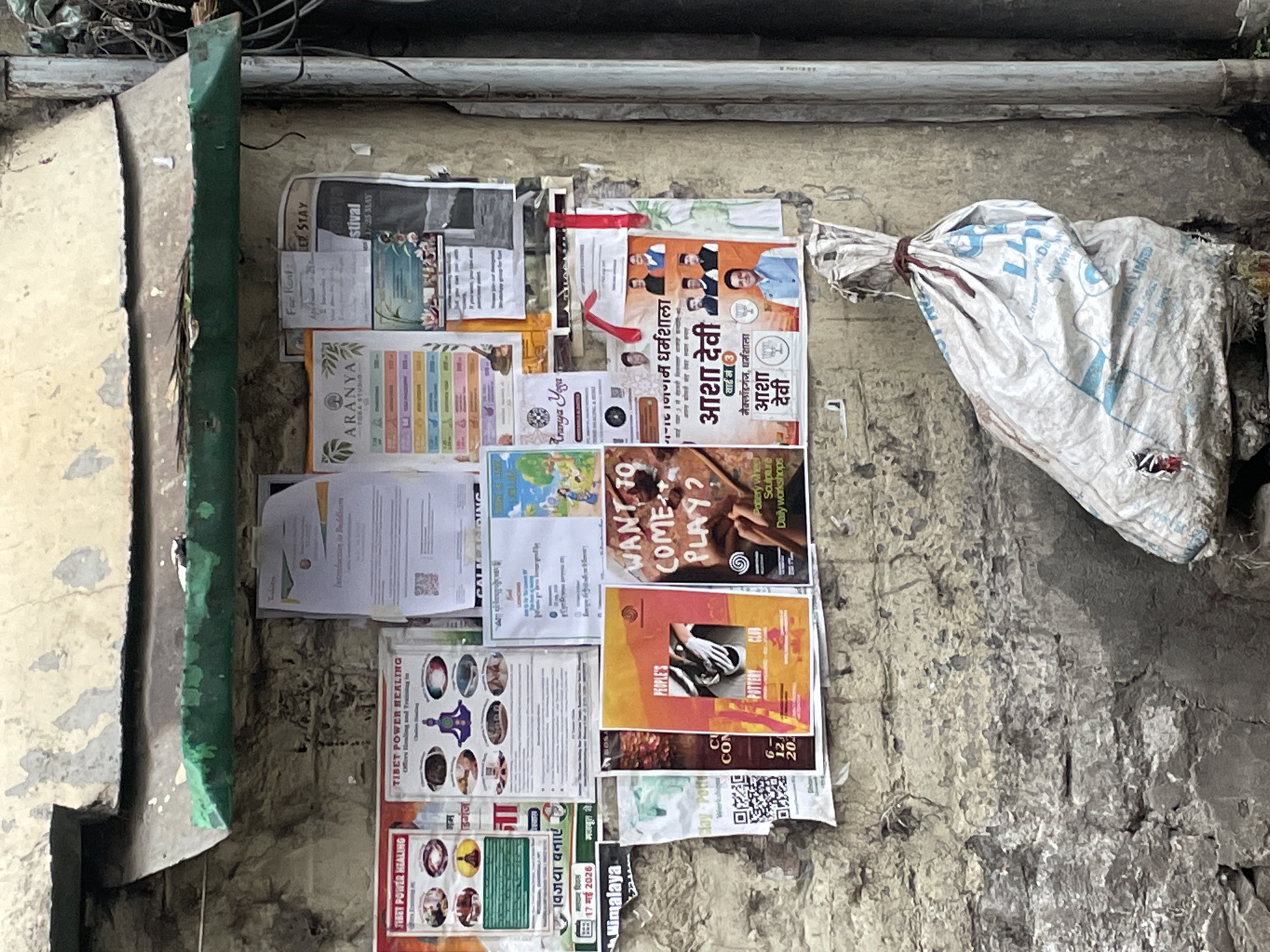 The image depicts a weathered exterior wall in Dharamshala, India, adorned with various posters and flyers. The wall itself is constructed of rough, textured material, showing signs of age and disrepair, with patches of peeling paint and mortar. A green-painted overhang extends from the top of the wall, providing some shelter.
Pinned to the wall are several notices, including advertisements for "Tibet Power Healing" and "Aranya Yoga." One poster features an introduction to Buddhism. Other flyers appear to be political campaign posters, displaying images of local politicians. The content of these posters suggests a mix of spiritual, wellness, and political themes common in the region.
In the foreground, a large, white, plastic bag, tied with twine, rests on the ground. The bag is partially filled and shows signs of wear, including a tear with some red material visible inside. To the right of the bag, a metal pipe, likely a drainpipe, runs vertically along the wall. The overall scene suggests an urban outdoor environment in a state of moderate disarray.